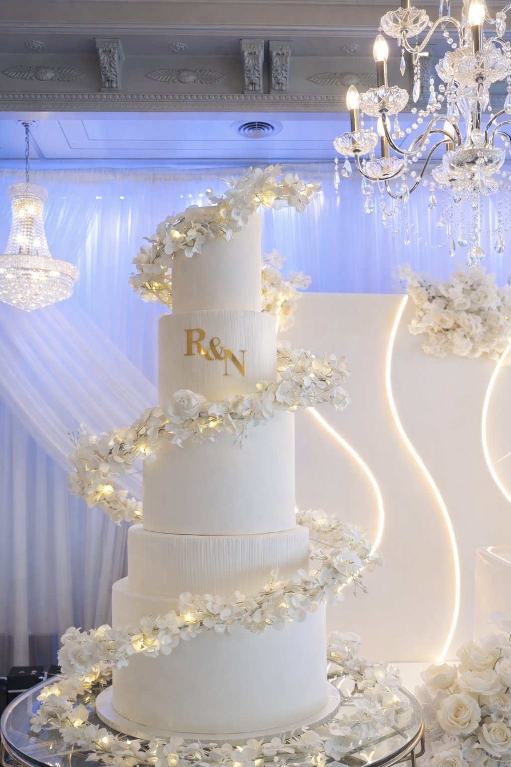 A tall, white wedding cake decorated with white flowers spiral all around, with a backdrop of elegant curtains, chandeliers, and floral arrangements, celebrating a special occasion.