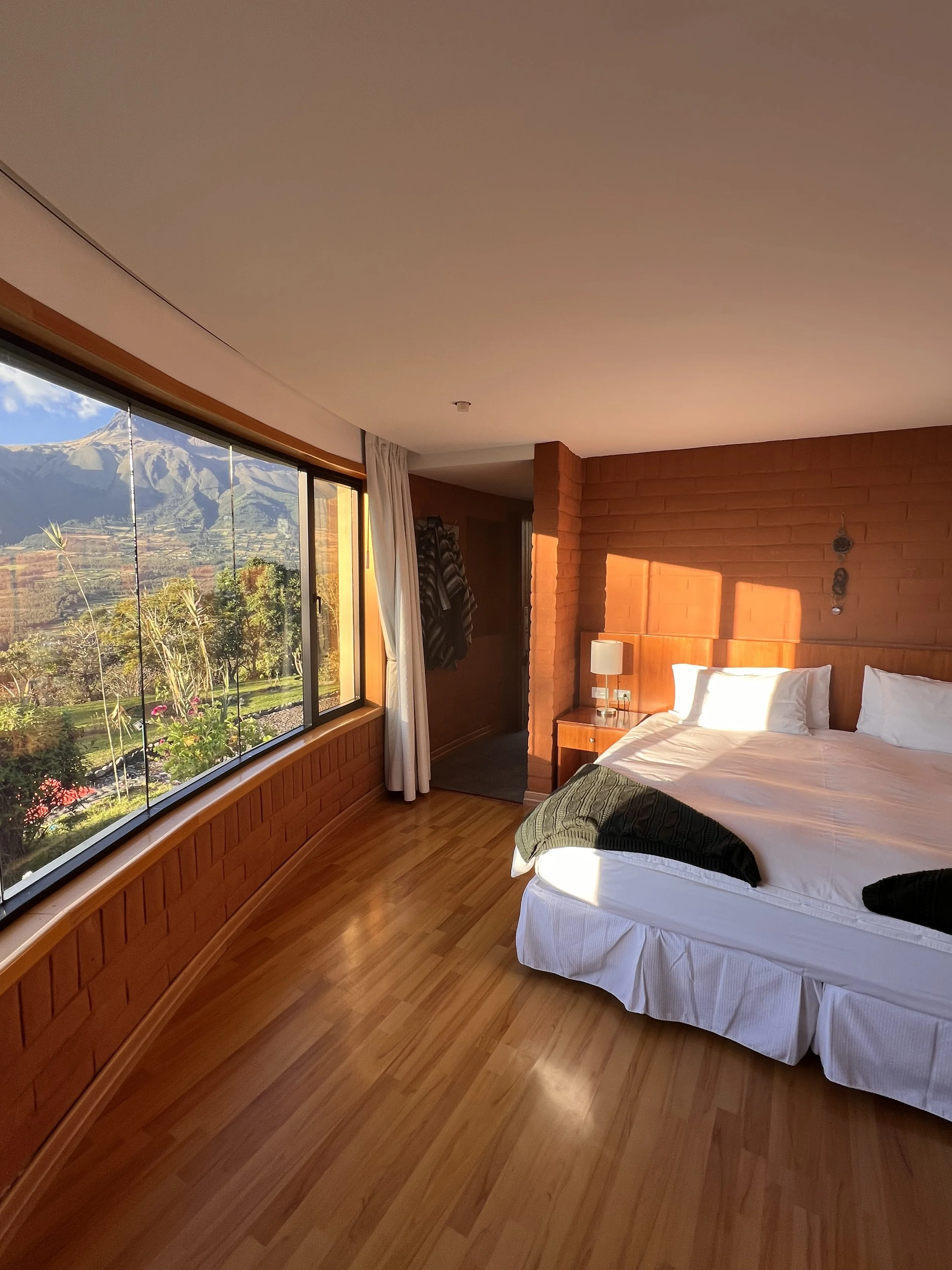 A hotel room with a large window showing a mountain range and landscape outside, a bed with white linens and a black sweater on it, a wooden nightstand with a lamp, and a brick wall behind the bed.