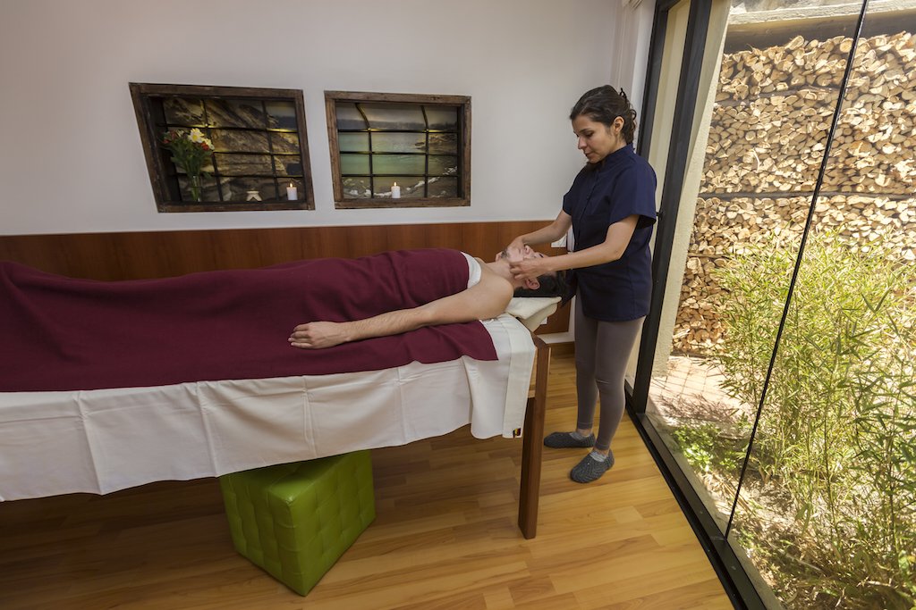 A woman giving a massage to a man lying on a massage table in a room with natural light, wooden floor, and a large window showing a garden with bamboo and stacked firewood.