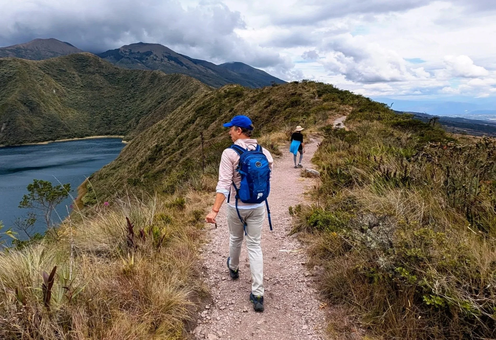 Hikers walking on a mountain trail alongside a lake with mountains in the background under a cloudy sky.