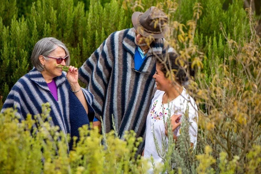 Three people smiling and chatting in a garden surrounded by tall green plants and bushes.