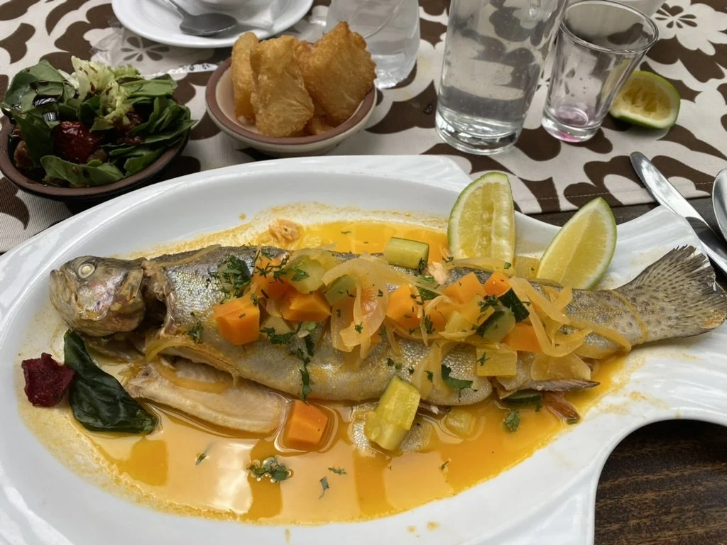 Whole cooked fish served on a white platter with lemon wedges, topped with vegetables and herbs, with side dishes including salad, fried food, water, and lemon slices on a dining table.