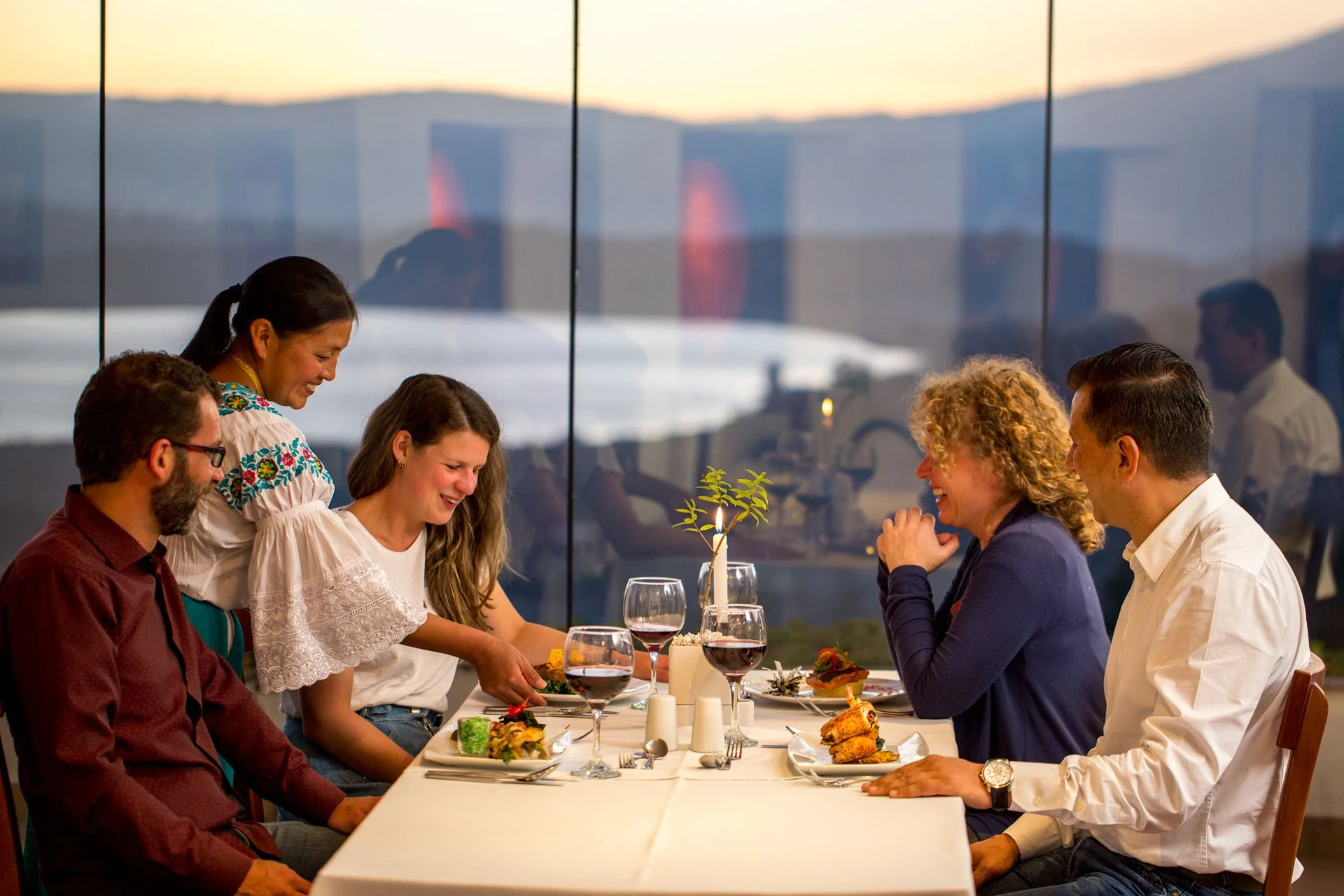 People dining at a restaurant table with a view of the mountains during sunset.