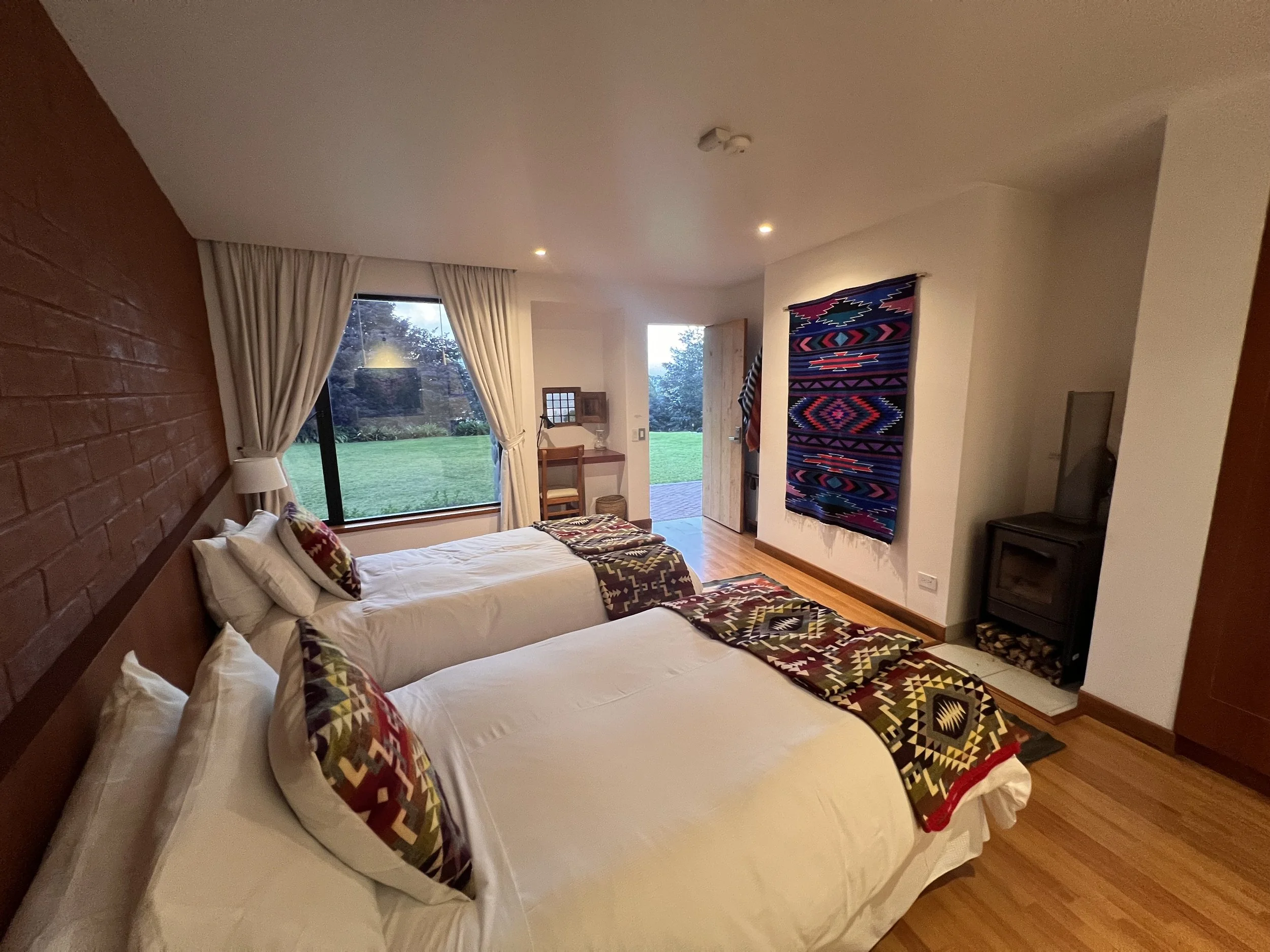 Interior of a bedroom with two beds, decorated with colorful patterned blankets and pillows, a large window with white curtains overlooking a green garden, a woven wall hanging, a small desk and chair near the door, a black television, and wood flooring.