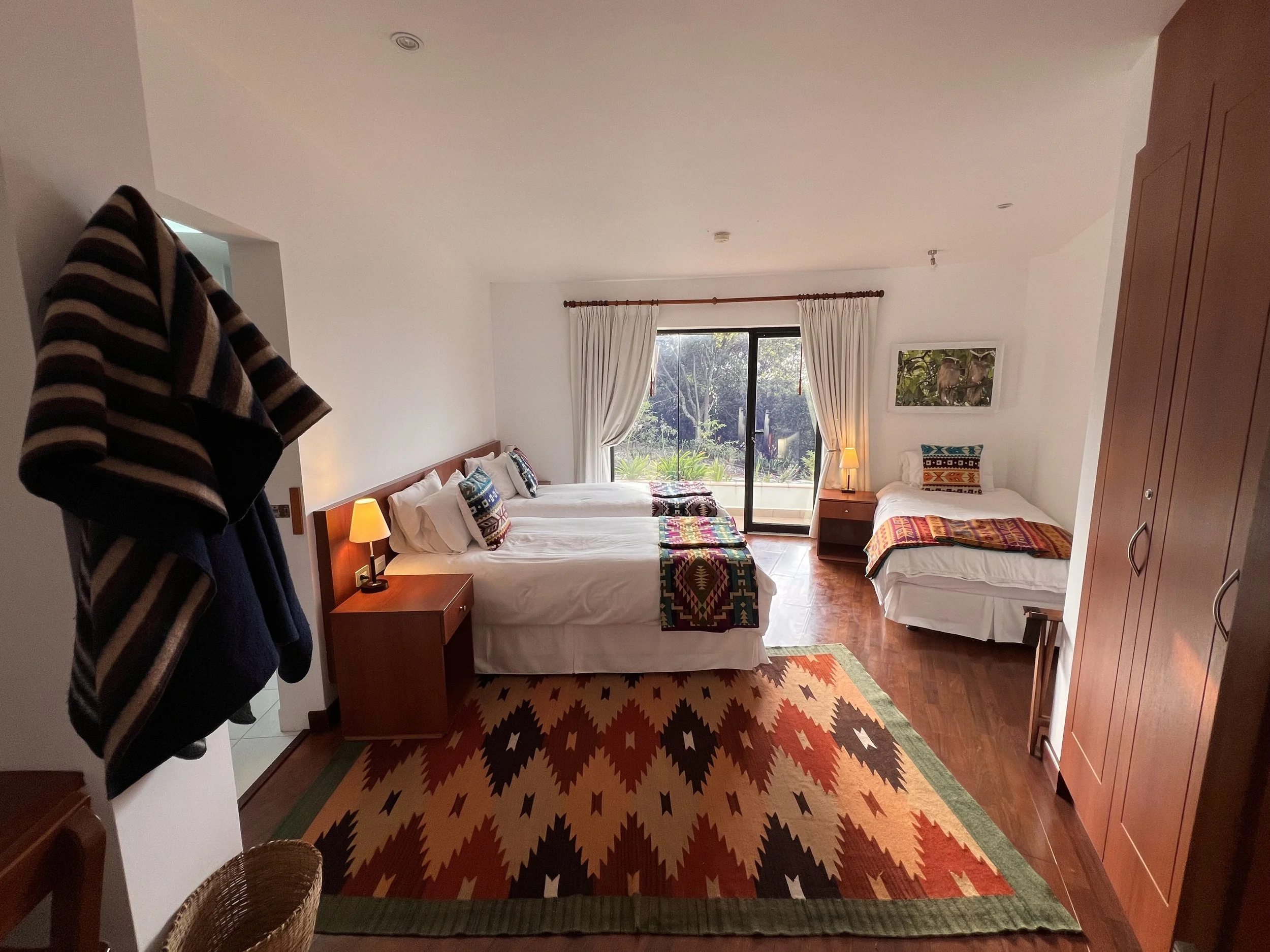 A bedroom with two beds, colorful patterned blankets, pillows, nightstands with lamps, large window with white curtains, and a colorful rug on a wooden floor. Part of a striped jacket is hanging on the left.