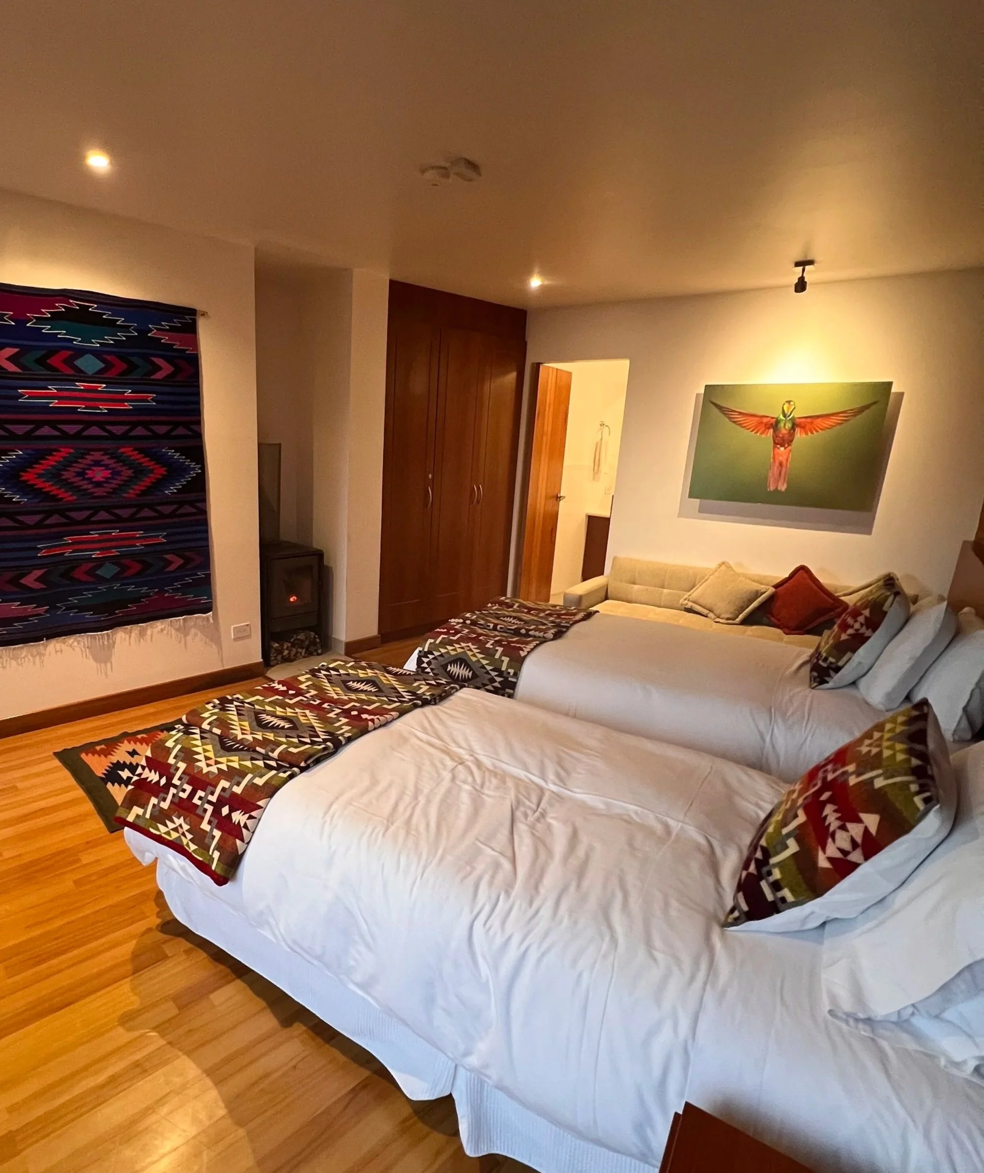 A bedroom with a white bed covered with a colorful tribal-patterned throw and pillows. Behind the bed, there are wooden cabinets, a mosaic wall hanging, a small black stand, and a framed artwork of a bird with outstretched wings. The room has hardwood floors and the ceiling has recessed lighting.