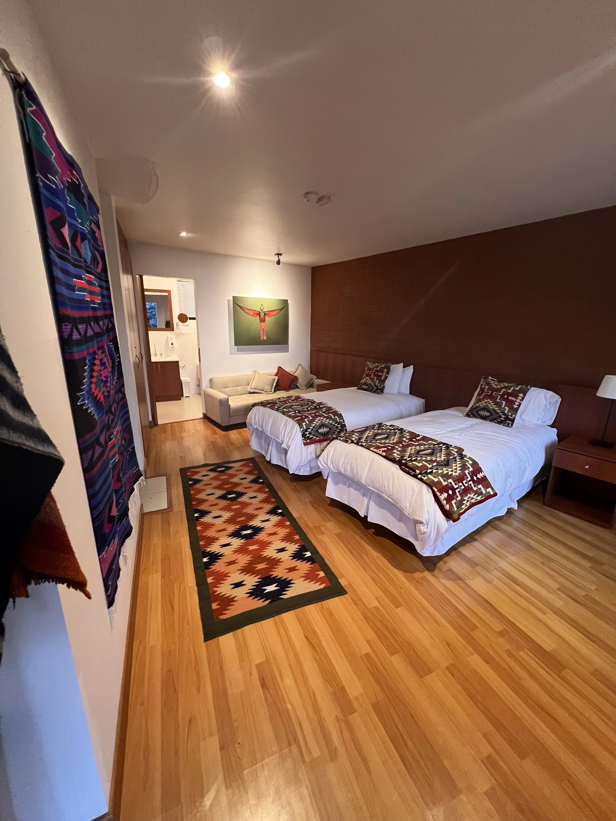 A hotel room with two beds, each with patterned bedding, a nightstand, a framed artwork of a bird with outstretched wings on the wall, a beige sofa with cushions, a colorful patterned rug on a wooden floor, a tapestry hanging on the wall, and a doorway leading to a bathroom.