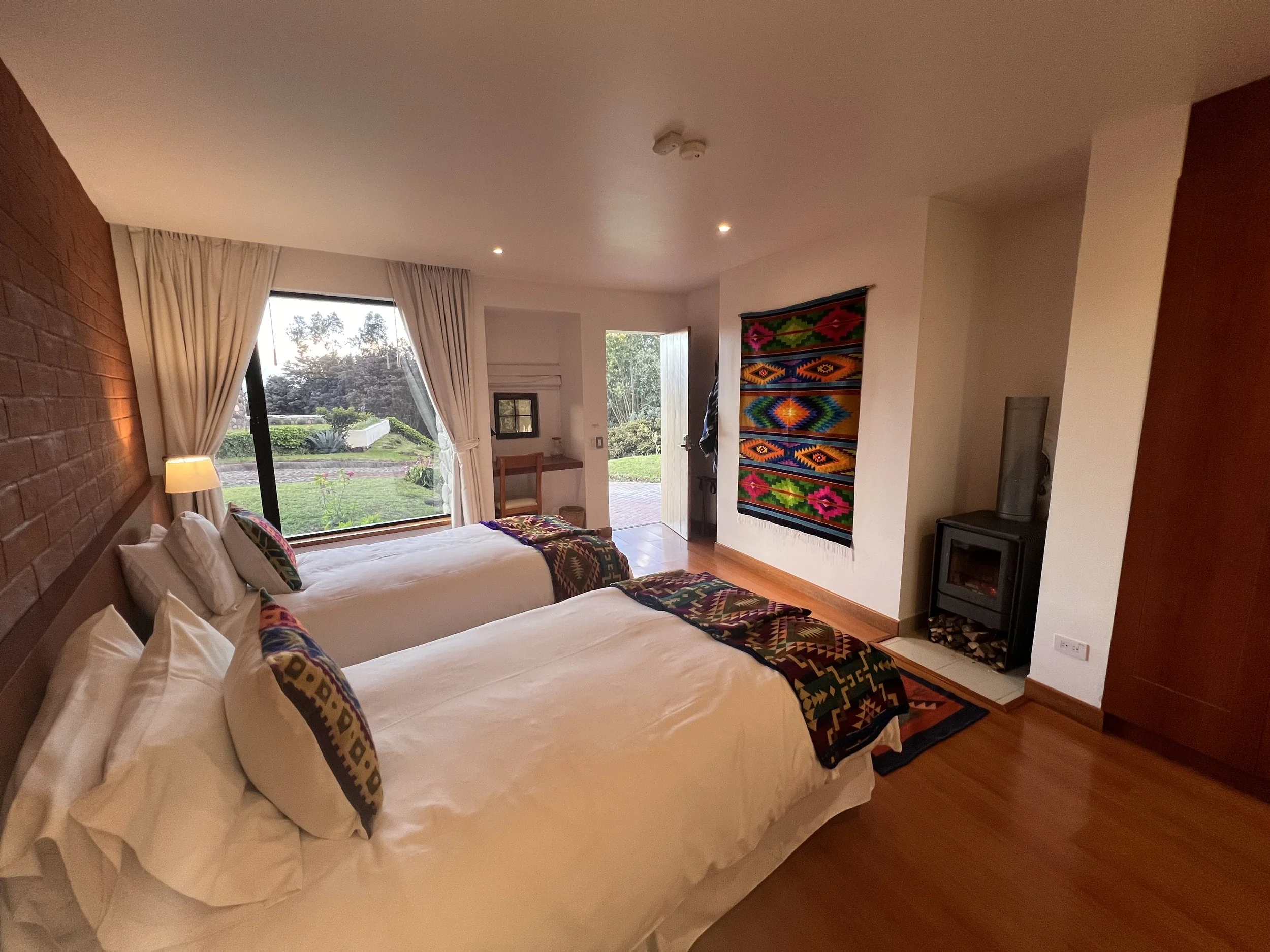 A cozy bedroom with two white beds, colorful patterned pillows, a large window with white curtains revealing a garden, and a wall hanging with a vibrant tribal pattern.