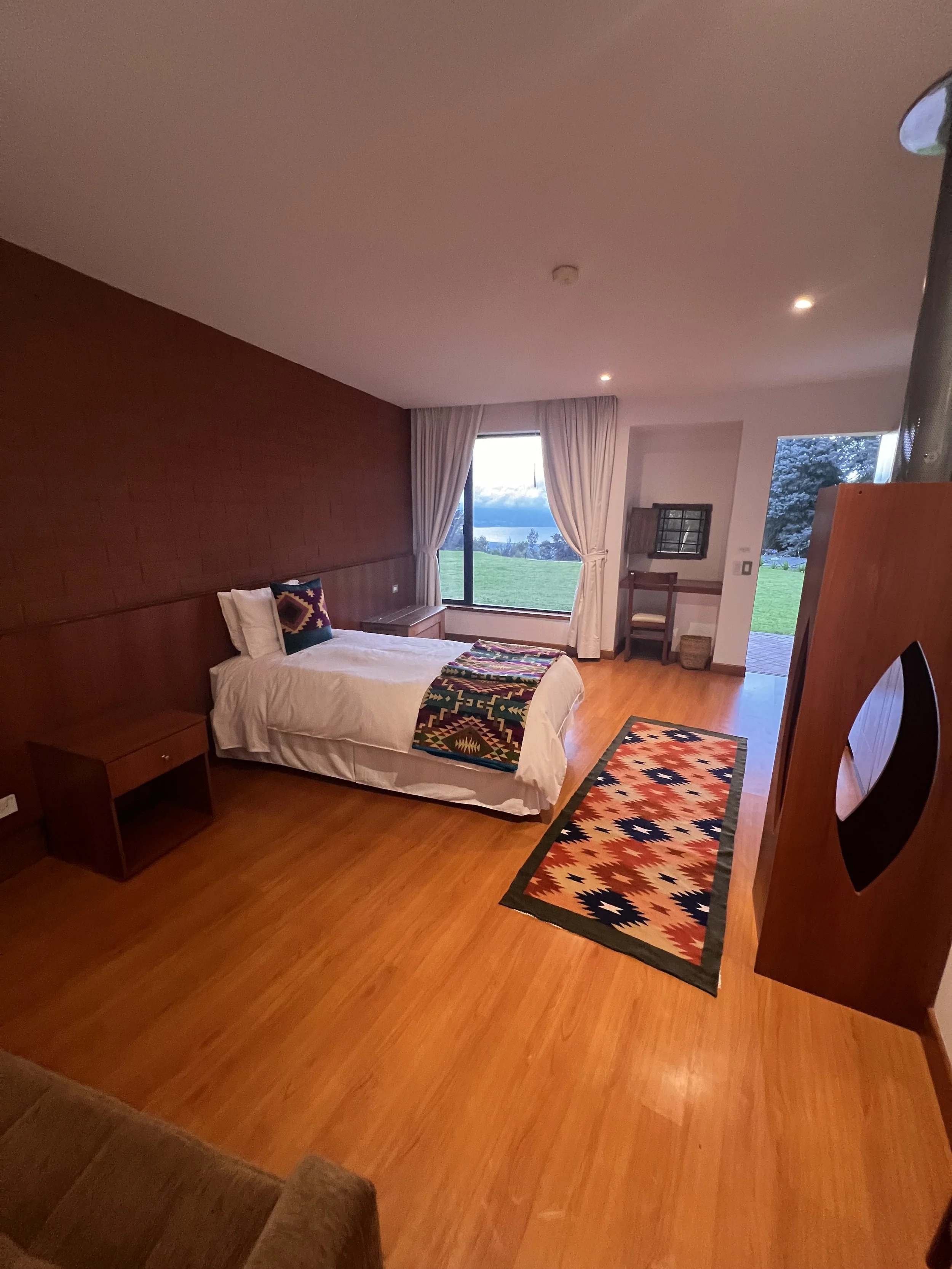 A cozy bedroom with a bed adorned with colorful pillows, a blanket, and a view of a lush green landscape through large windows, illuminated by warm natural light and ceiling spotlights.