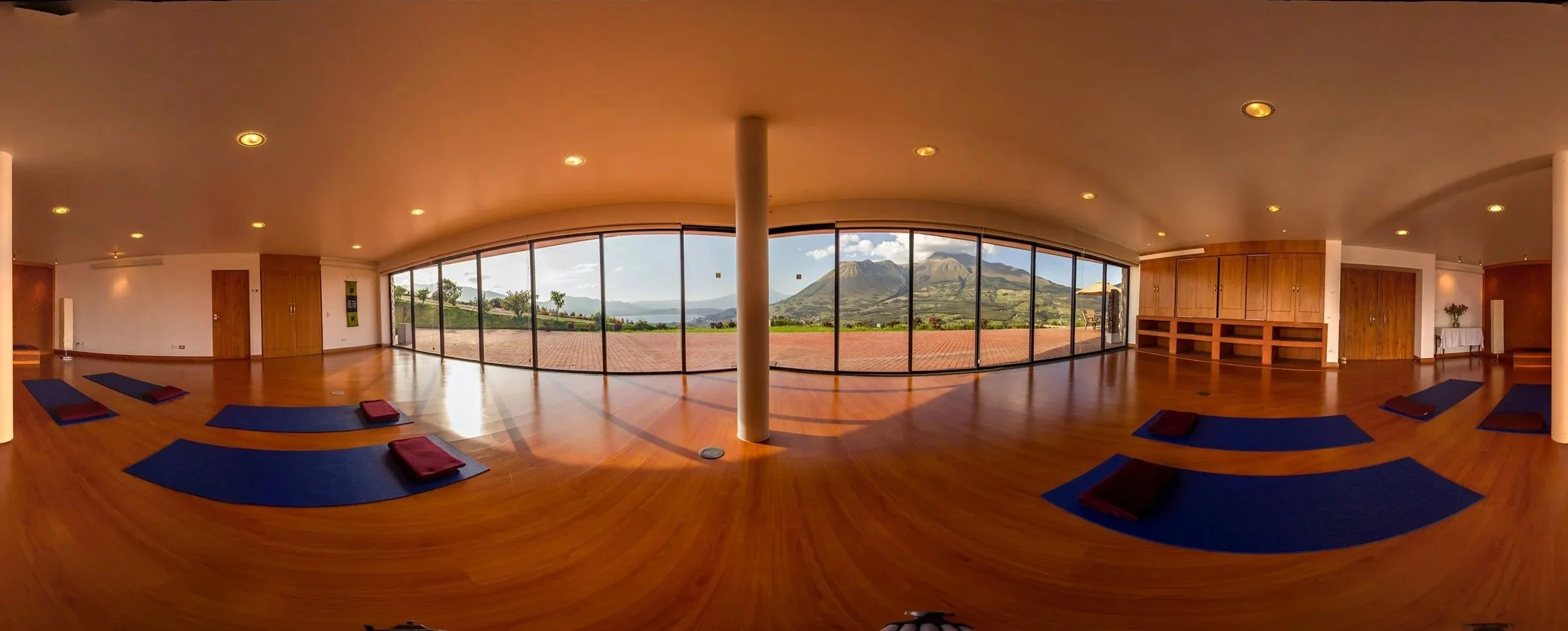 Interior view of a yoga studio with blue mats and folded towels, large windows overlooking mountains and a cloudy sky, wooden floors and minimal decor.