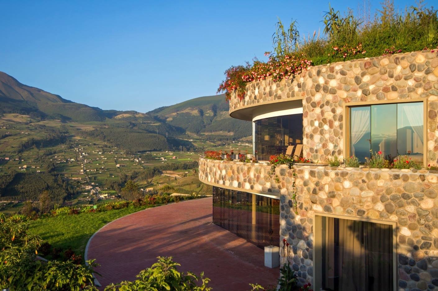 A modern stone house with curved balconies and large windows, overlooking a mountainous landscape under a clear blue sky.