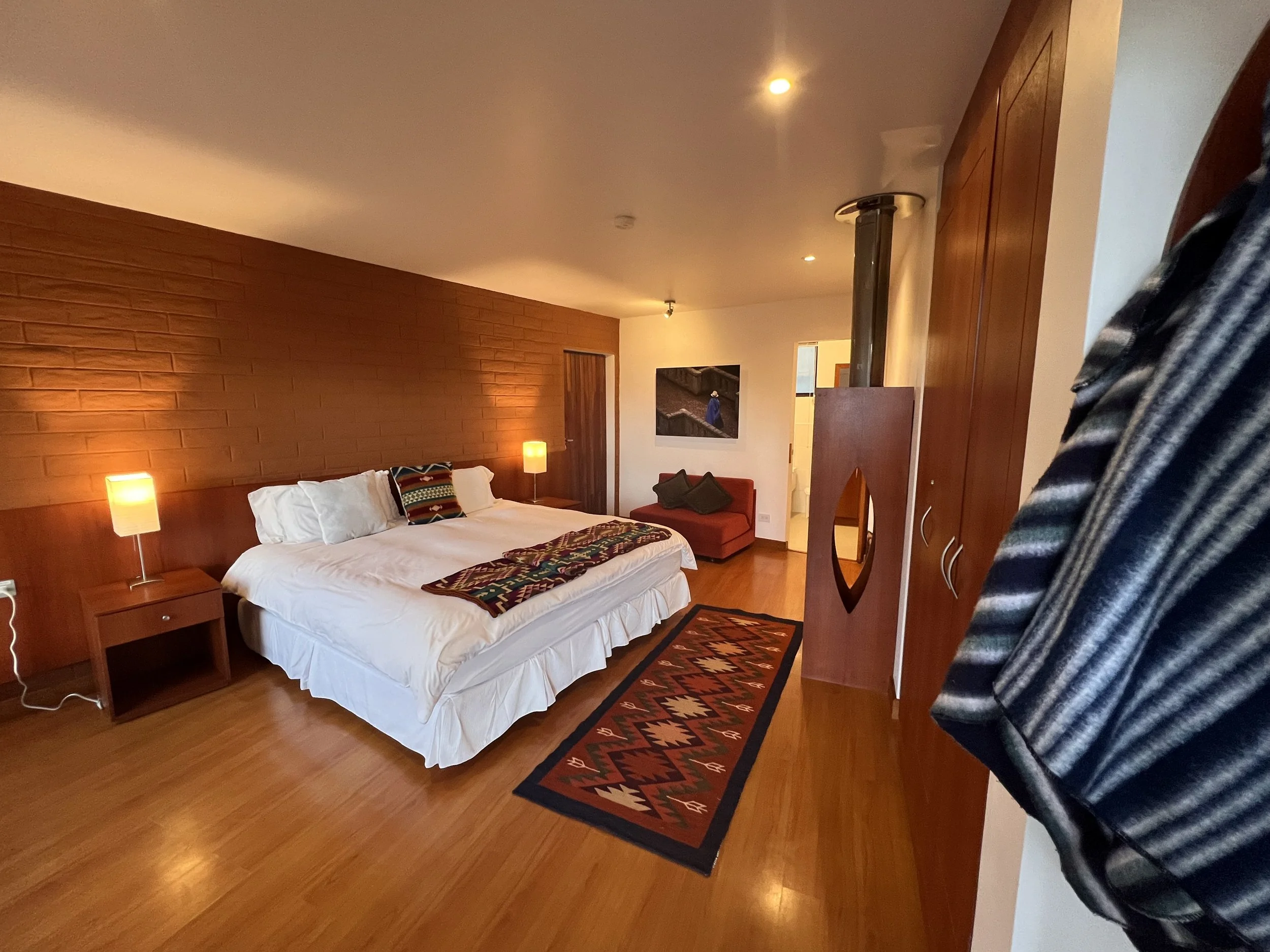 A hotel room with a bed, two bedside lamps, a patterned throw on the bed, a chair, a framed picture, a wooden wardrobe, and a hanging quilted blanket.