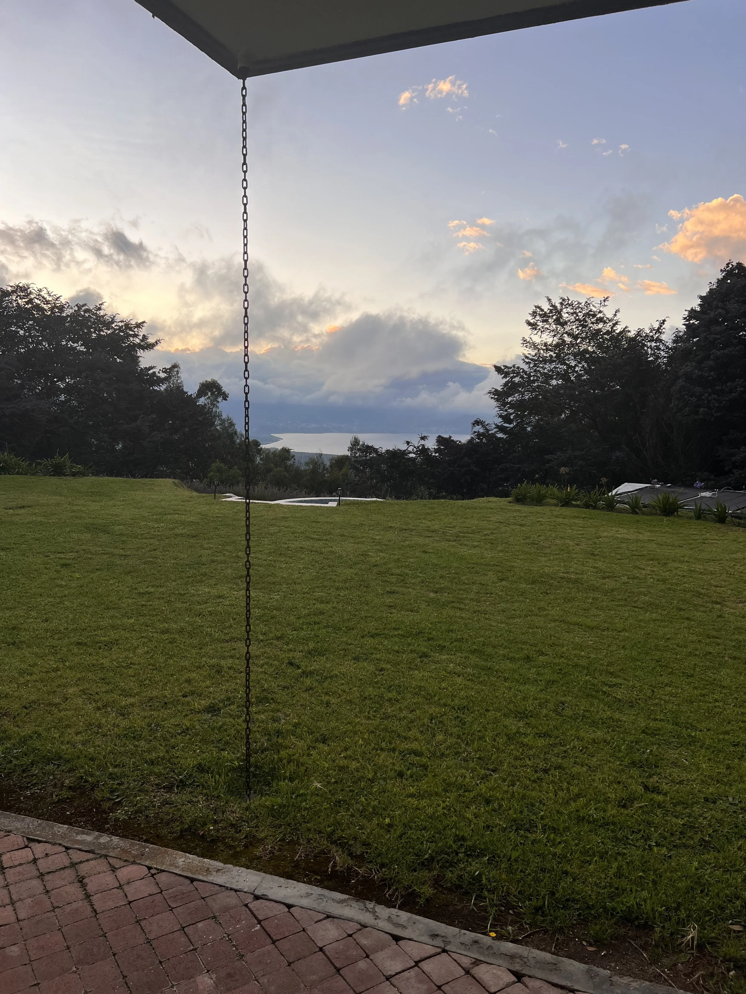 A backyard with green grass, trees, and a view of mountains and a body of water visible in the distance at sunset, with a porch or balcony and a chain hanging from the ceiling.