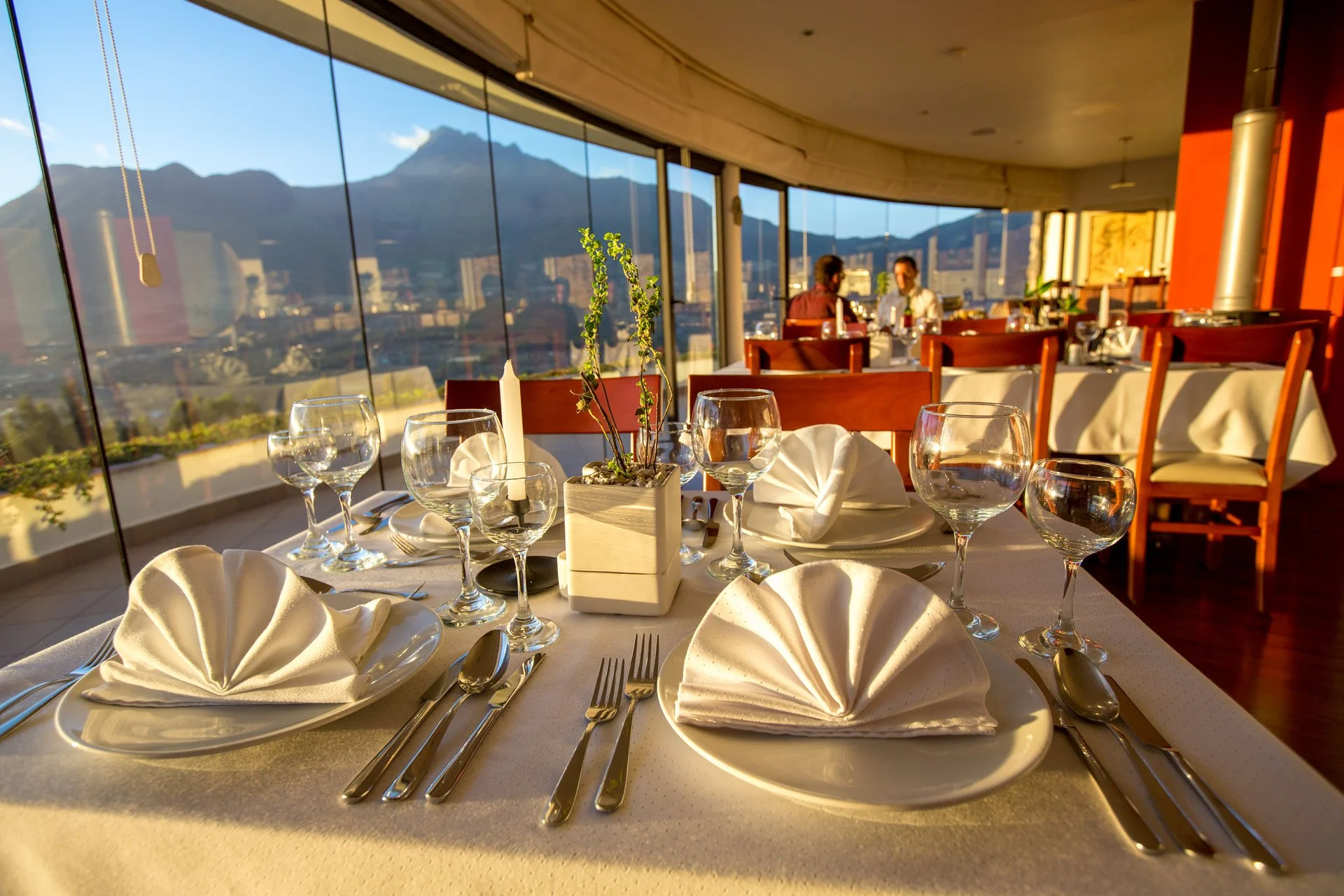 Elegant dining table set with wine glasses, plates with folded napkins, and silverware in a restaurant with large windows showing mountains and cityscape during sunset.