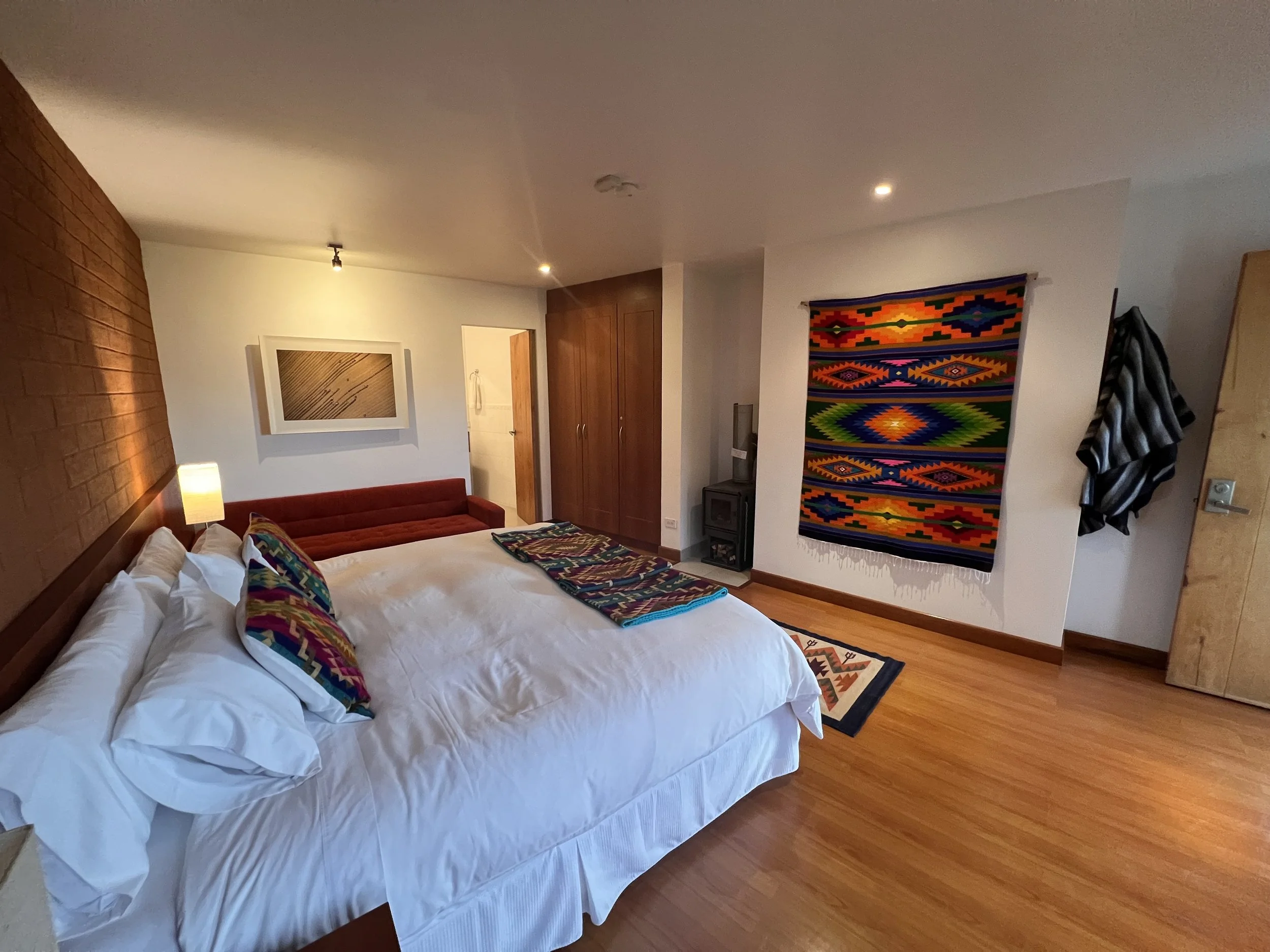 A bedroom with white and brick walls, a wooden floor, a bed with white linens and colorful pillows, a red sofa, framed artwork, colorful woven wall hangings, and various furniture and decorations.