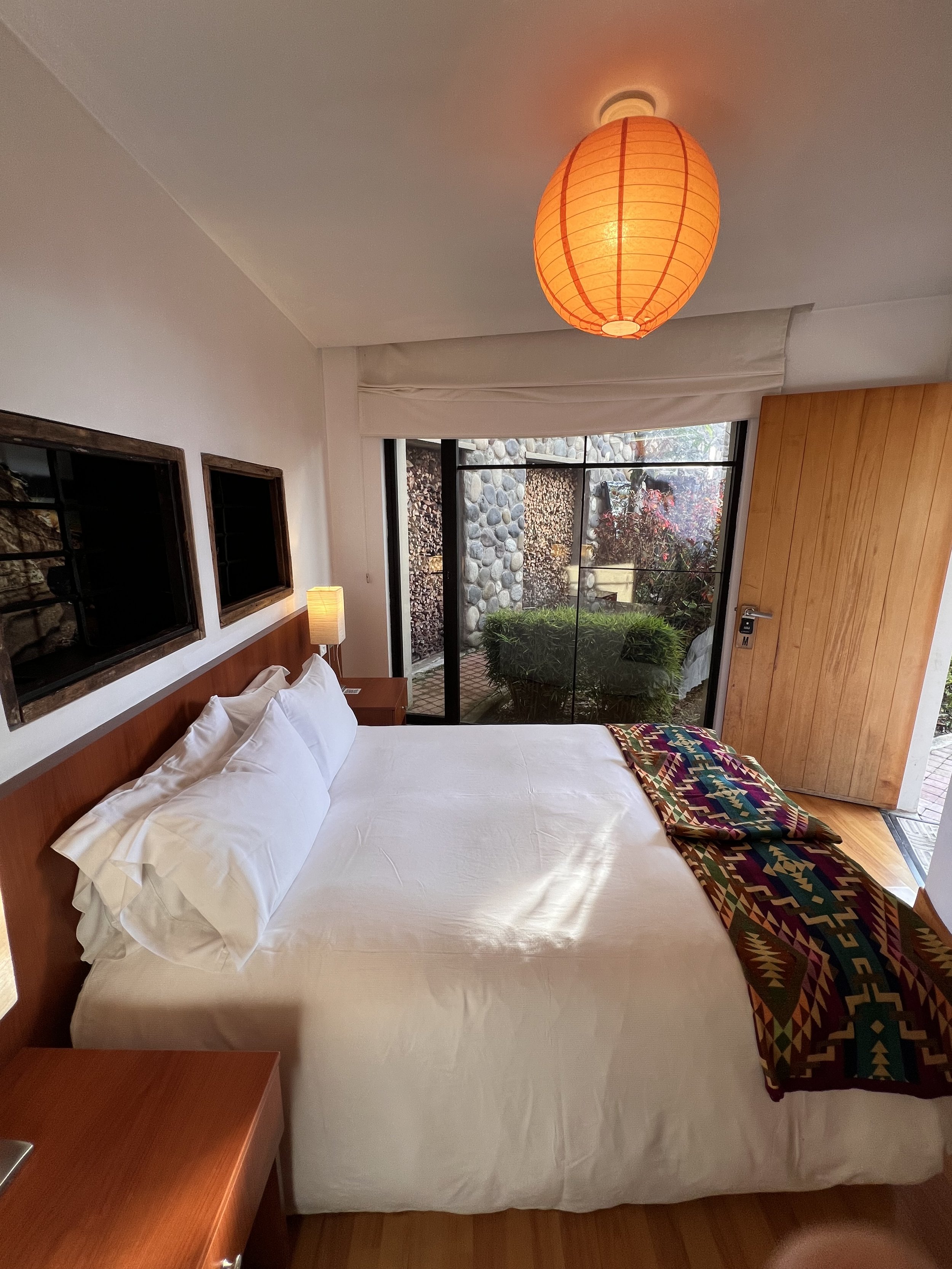 A bedroom with a bed made with white sheets and pillows, a colorful woven blanket at the foot, a wooden headboard, a bedside table with a lamp, large window revealing plants outside, and an orange paper lantern ceiling light.