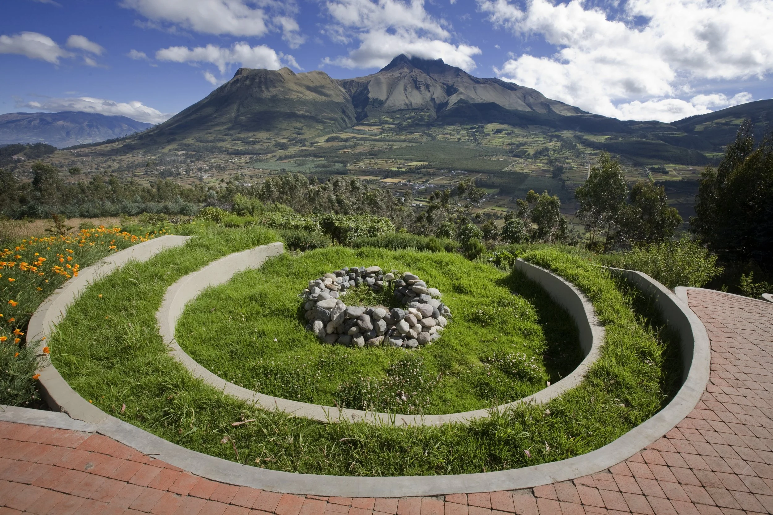 A landscaped garden with a circular flower bed, in front of a mountainous landscape with green fields and blue sky with clouds.