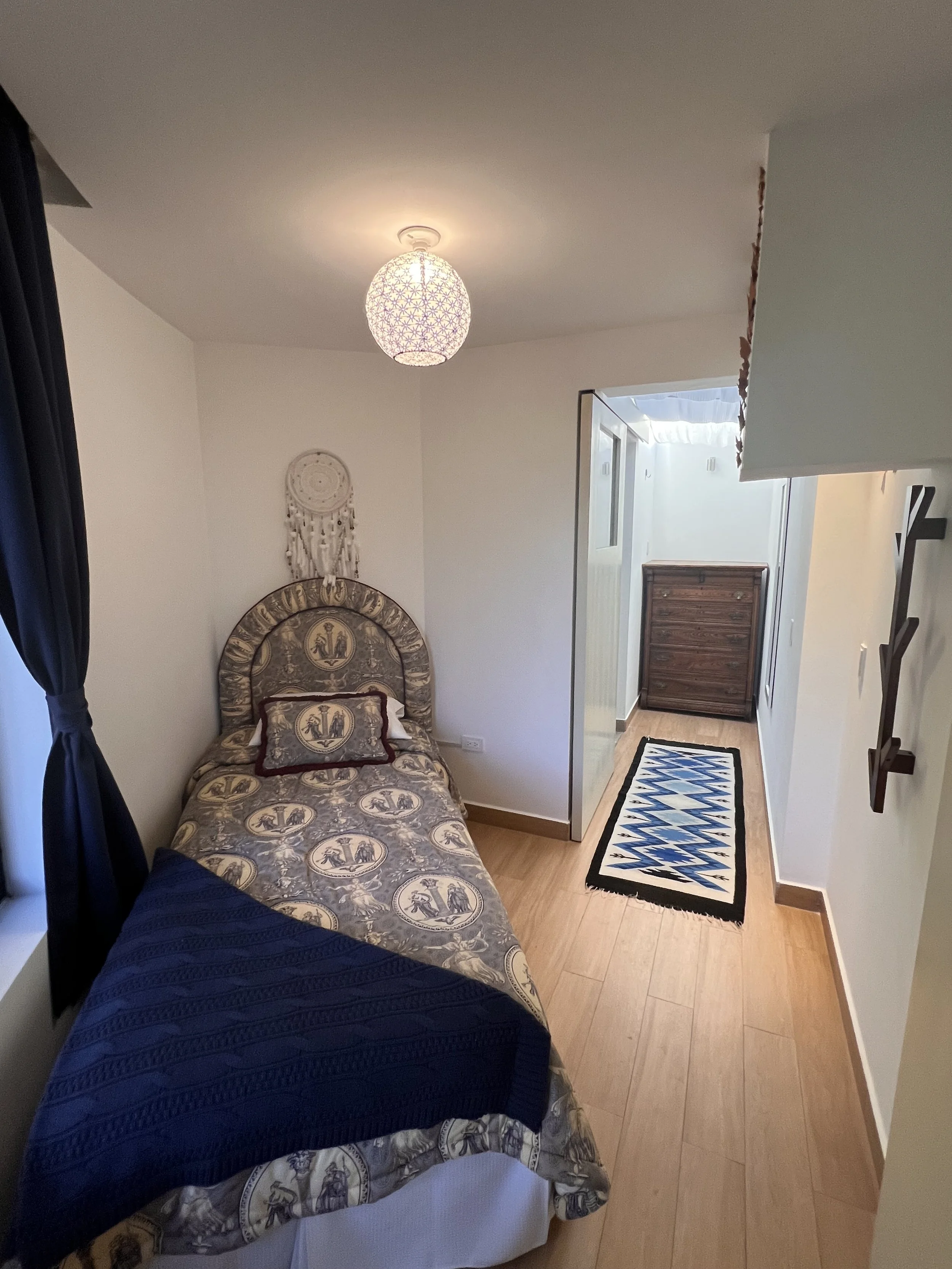 Small bedroom with a bed covered with a patterned bedspread featuring ancient Greek scenes and a decorative pillow. A dreamcatcher hangs on the wall above the headboard. There's a nightstand or a piece of furniture visible at the foot of the bed. The room has light-colored walls and a wooden floor. A framed patterned rug is on the floor in front of a hallway leading to other rooms, with a chest of drawers visible in the hallway. A hanging ceiling light is switched on, illuminating the room.