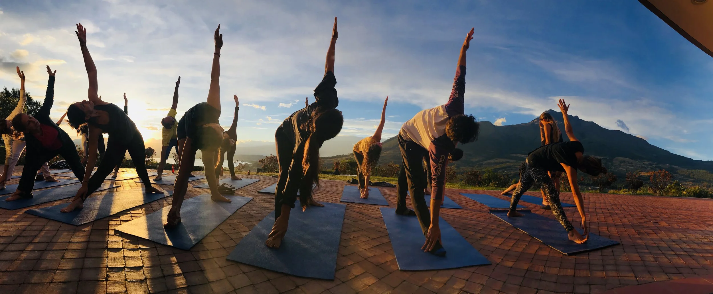 Group of people practicing yoga outdoors on mats during sunset, with a mountain view in the background.