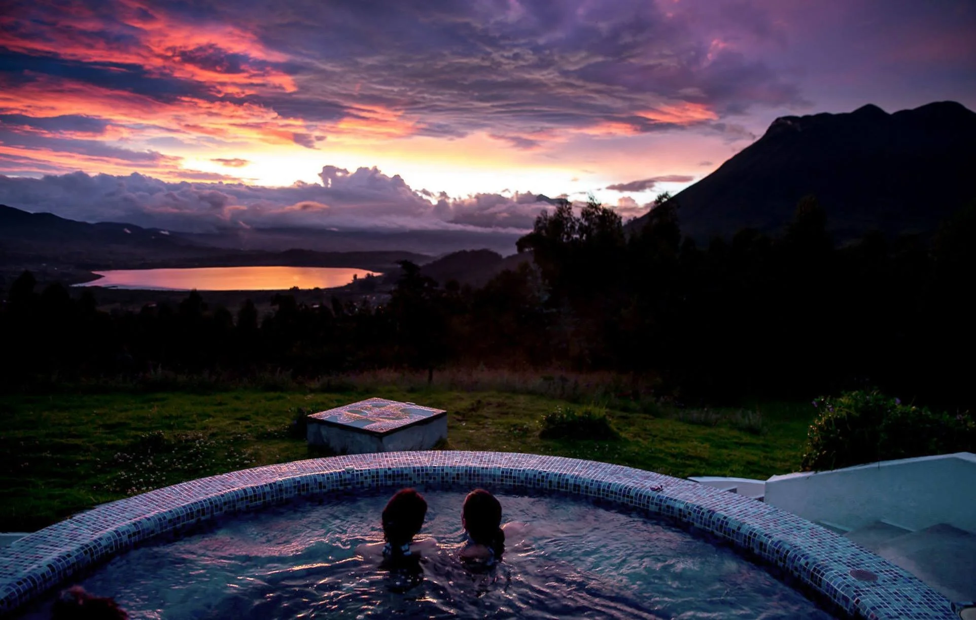 Two children sitting in a hot tub outdoors, with a scenic backdrop of a lake, mountains, and a colorful sunset sky with clouds.