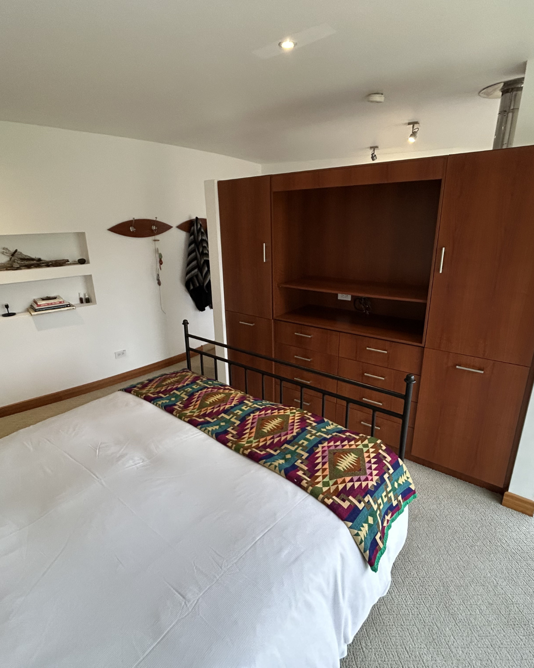 A bedroom with a bed covered in white bedding and a colorful, patterned blanket at the foot. A large wooden entertainment cabinet is against the wall, with open space for a TV and multiple drawers. To the left, a small wall shelf with books and decorative items, and a coat hanging on the wall. The ceiling has recessed lighting and a small vent pipe.