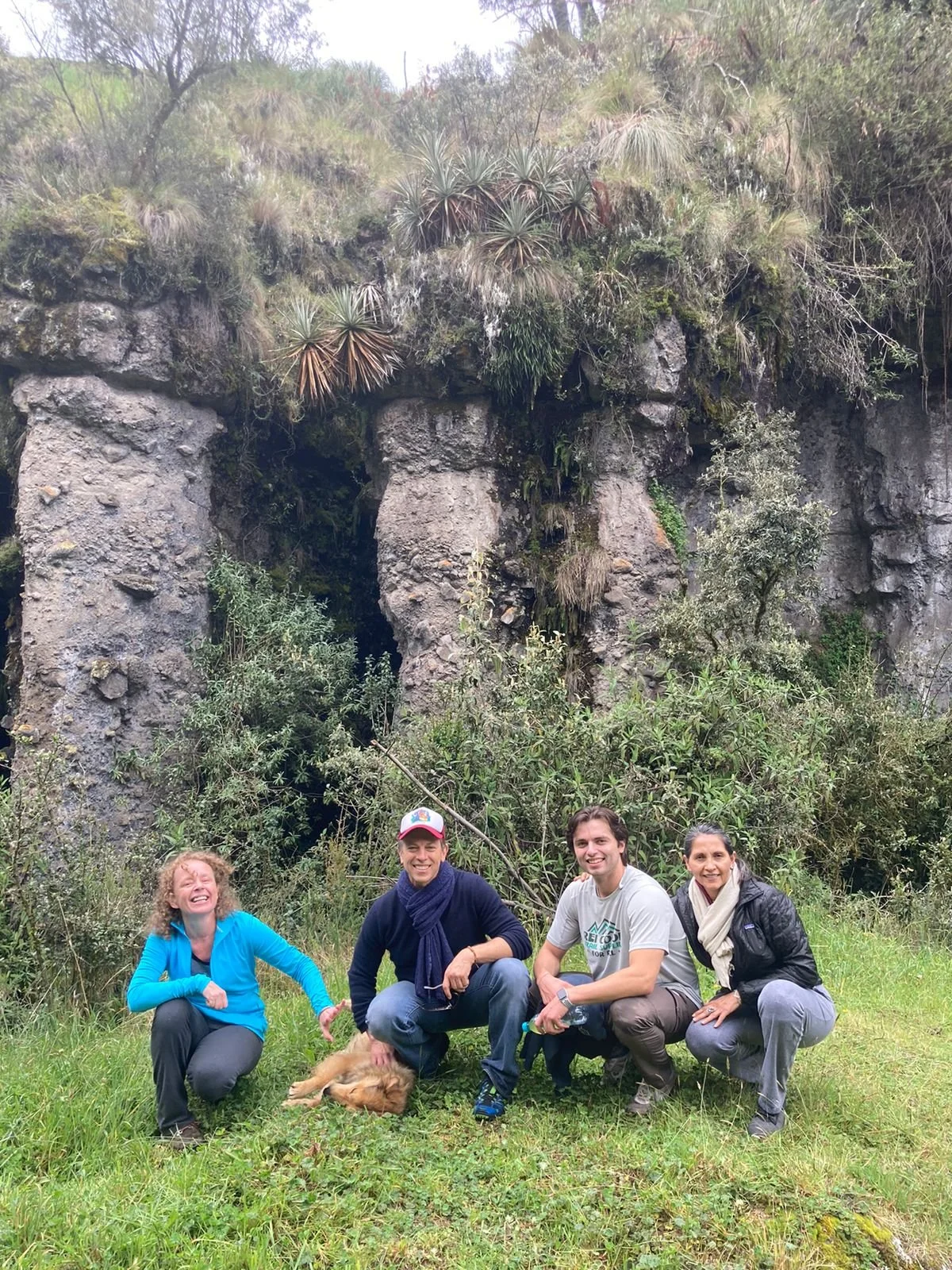 Four people smiling and posing outdoors in front of a rocky, plant-covered hillside with a small cave or hollow in the rocks, along with some trees and bushes, during daytime.