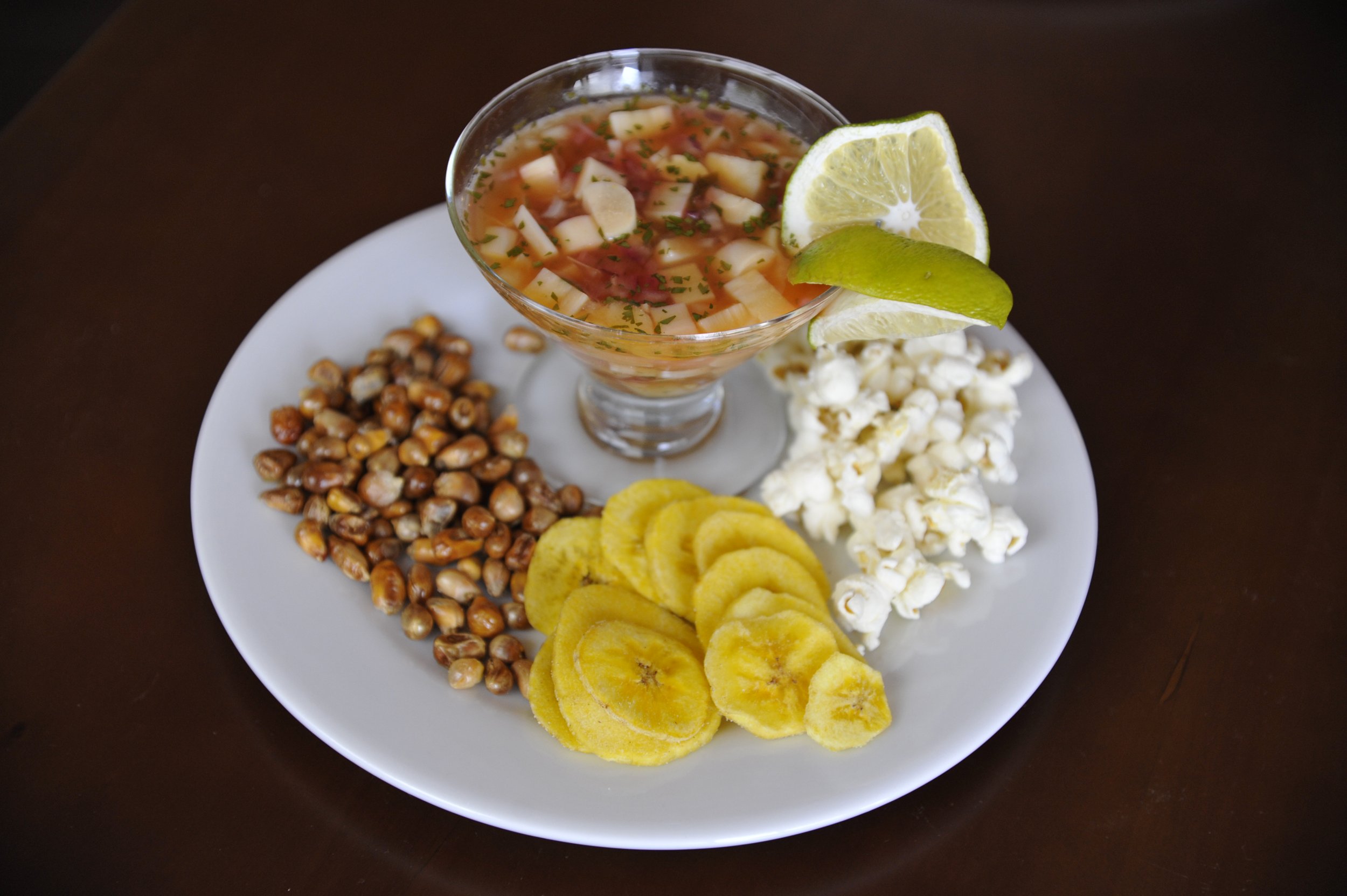 A white plate with sliced bananas, popcorn, roasted corn, and potato chips, topped with a glass of cold fruit cocktail garnished with lemon and lime wedges.