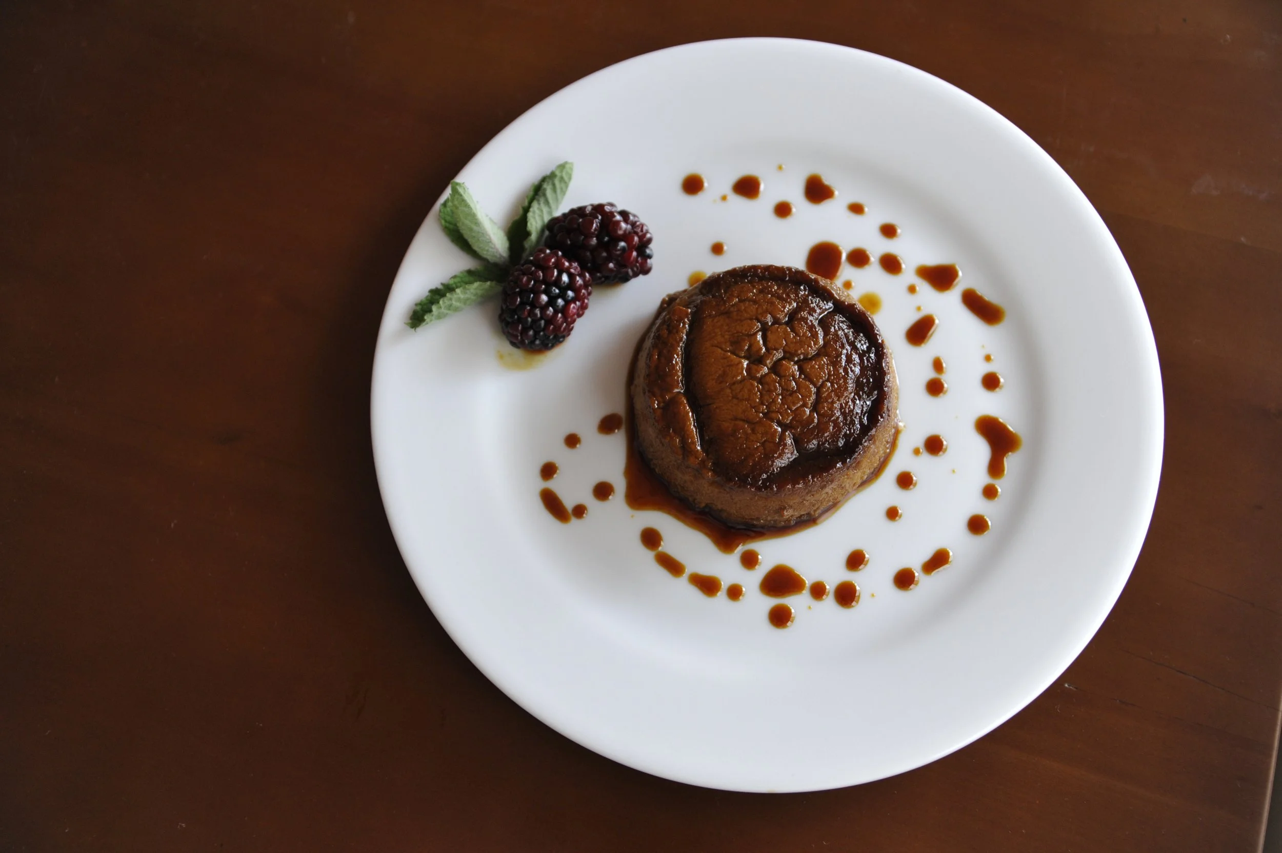 A white plate with a baked chocolate dessert, garnished with two blackberries and fresh mint leaves, and drizzled with caramel sauce, on a dark wooden table.