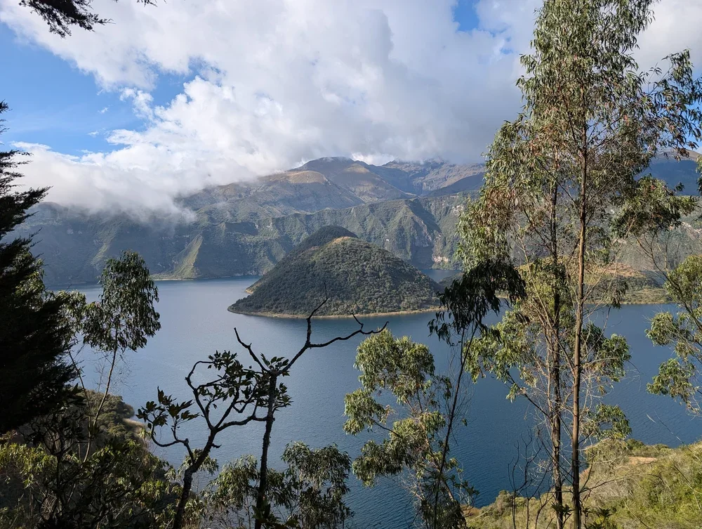 A scenic view of a lake surrounded by green mountains and trees, with partly cloudy skies overhead.