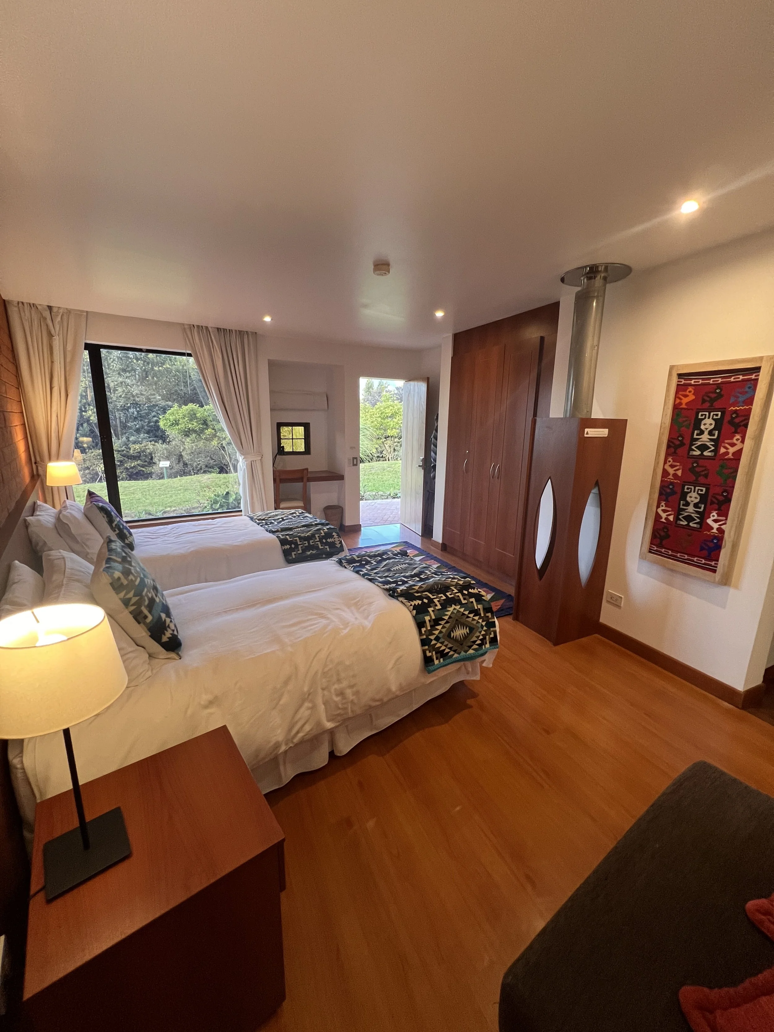 A cozy bedroom with two white beds with patterned pillows and blankets, a wooden nightstand with a lamp, large window with white curtains revealing greenery outside, a wooden wardrobe, a small sitting area with a desk, and colorful artwork on the wall.