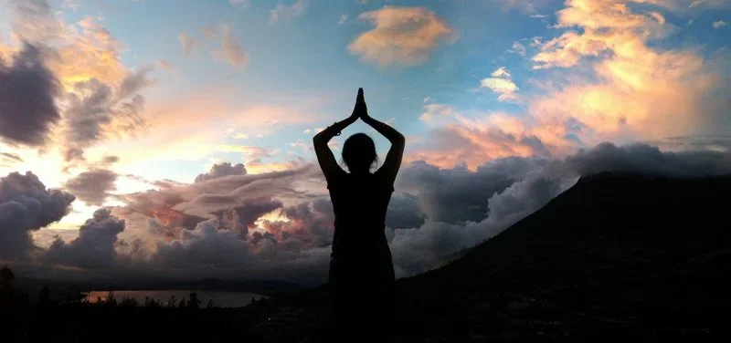 Person practicing yoga outdoors during sunset with clouds and mountain landscape in background.