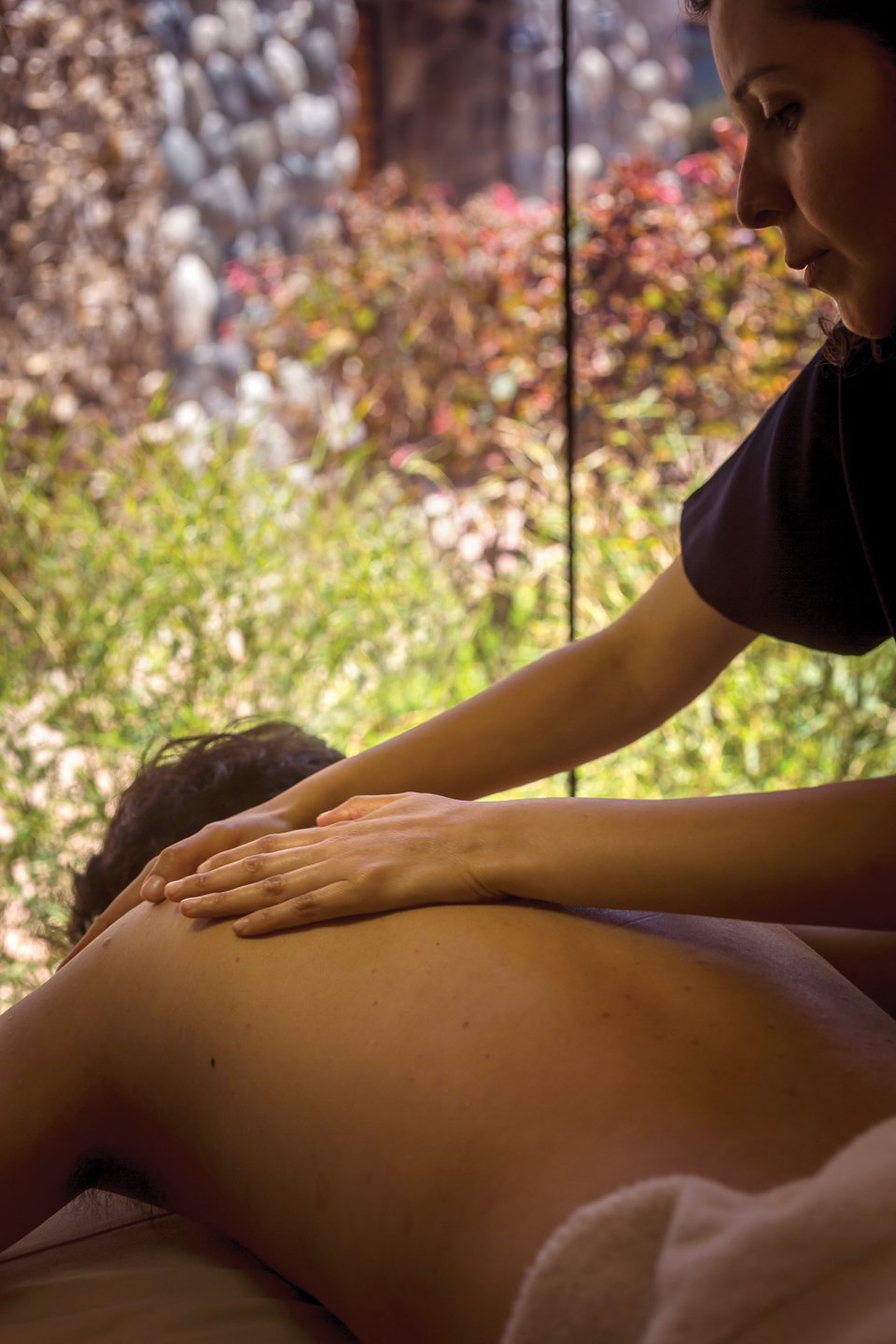 A person receiving a massage, lying face down with a therapist applying pressure on their back, in a serene setting with blurred natural background.