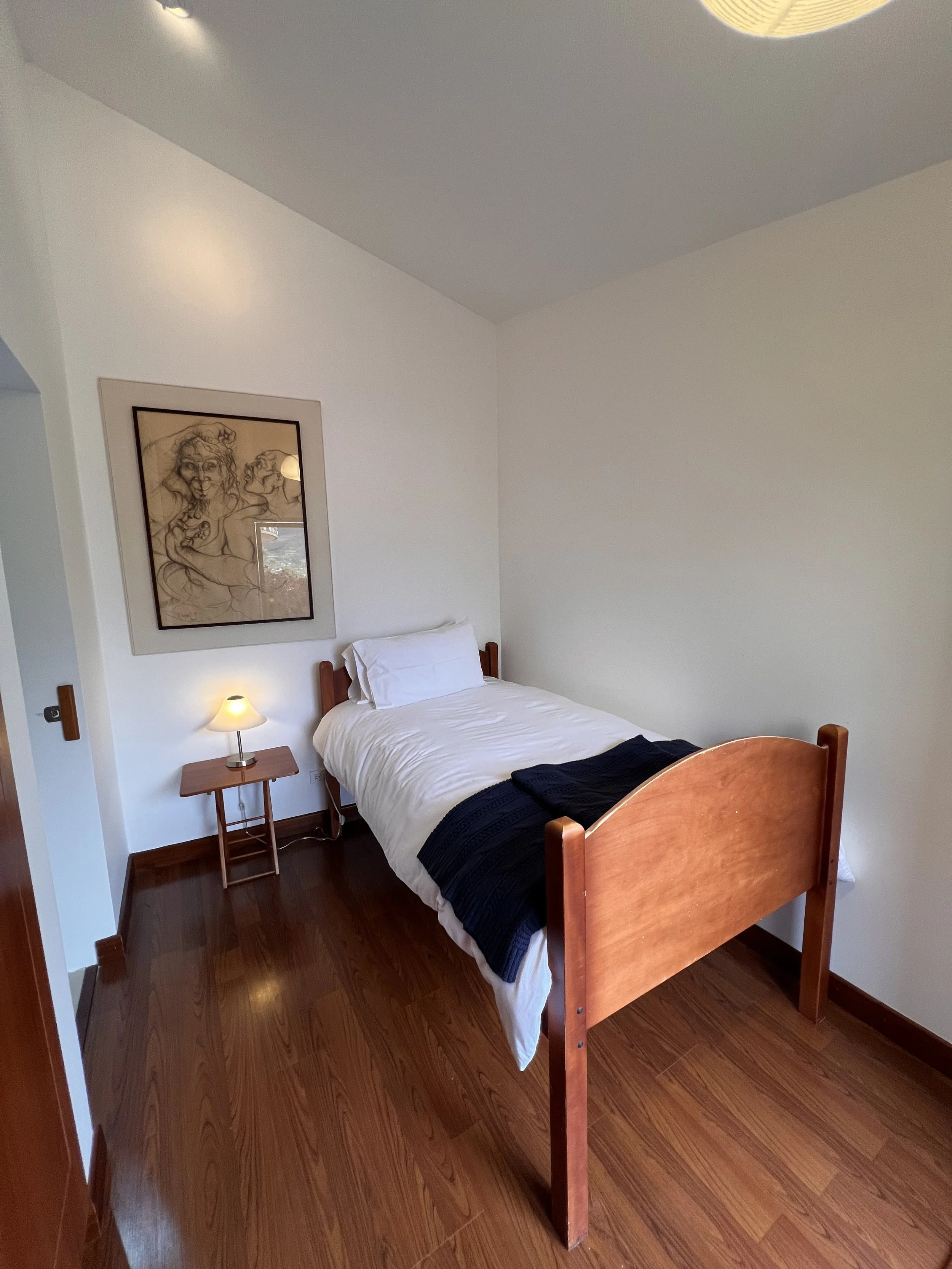 A bedroom with a wooden bed, white bedding, a black blanket at the foot, a small wooden nightstand with a lamp, a framed sketch of women on the wall, and hardwood floors.