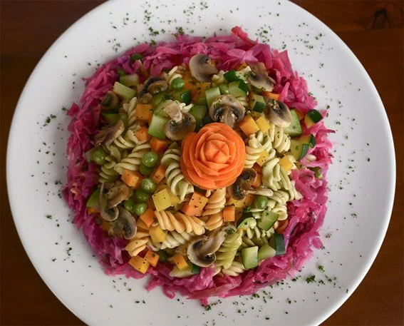 A plate of colorful pasta salad with chopped vegetables, mushrooms, and cheese, garnished with a carved carrot in the shape of a flower and pink shredded cabbage around the edges.