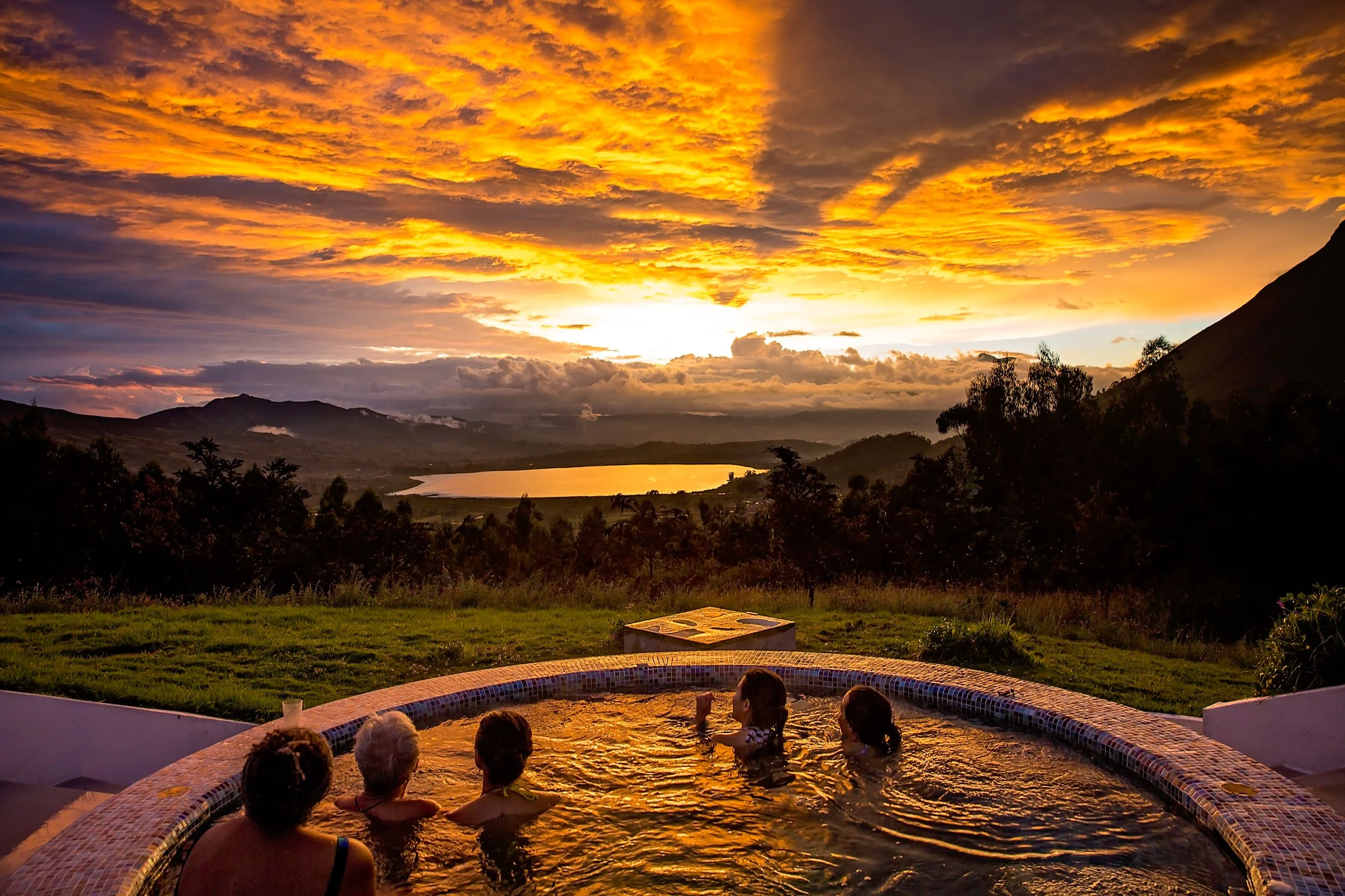 People relaxing in a hot tub during sunset with a scenic view of mountains, a lake, and a colorful sky filled with orange and yellow clouds.