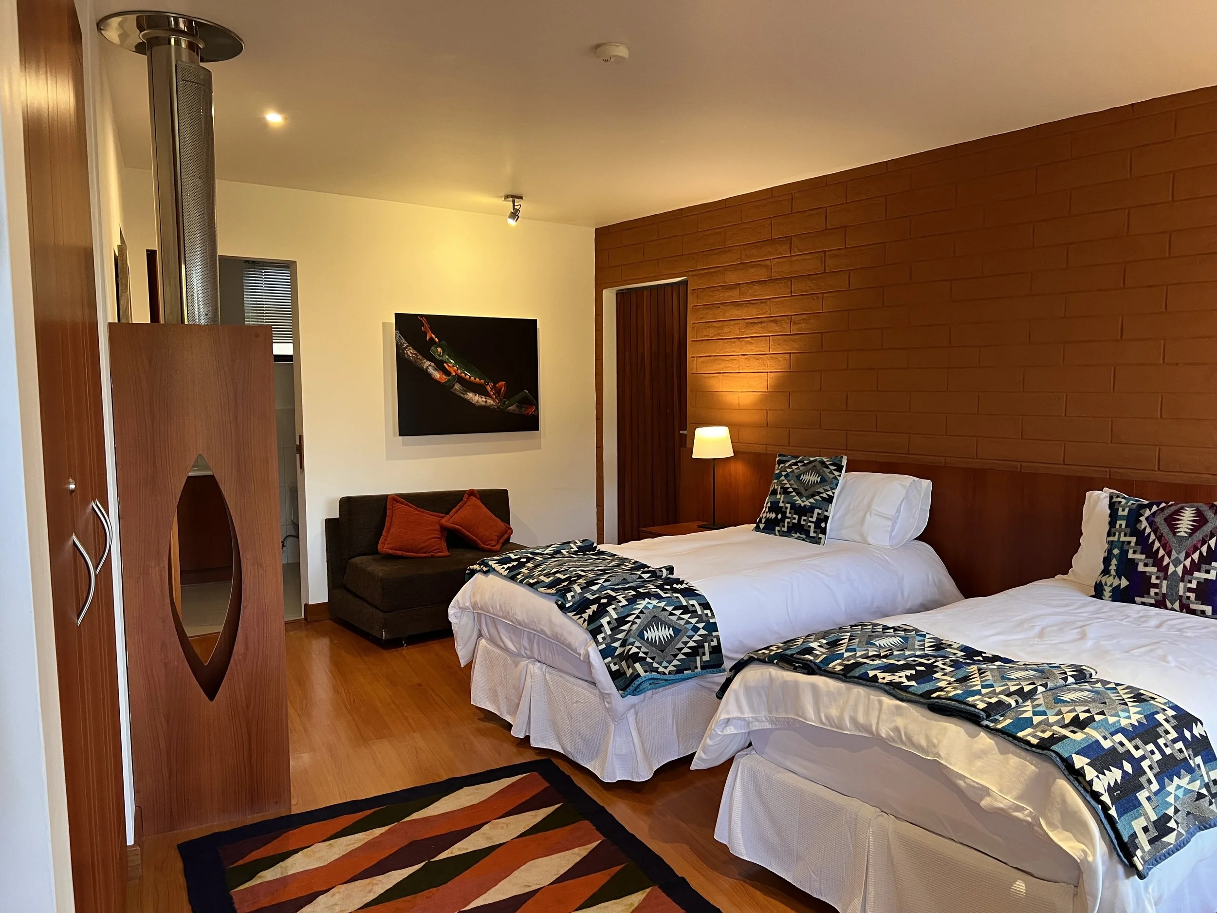 A cozy bedroom with two twin beds dressed in white linens, patterned blankets, and pillows. There is a wooden headboard, a standing lamp, a framed picture of a colorful lizard or frog on the wall, and a dark sofa with orange pillows. The room has wooden flooring, a brick accent wall, and warm lighting.