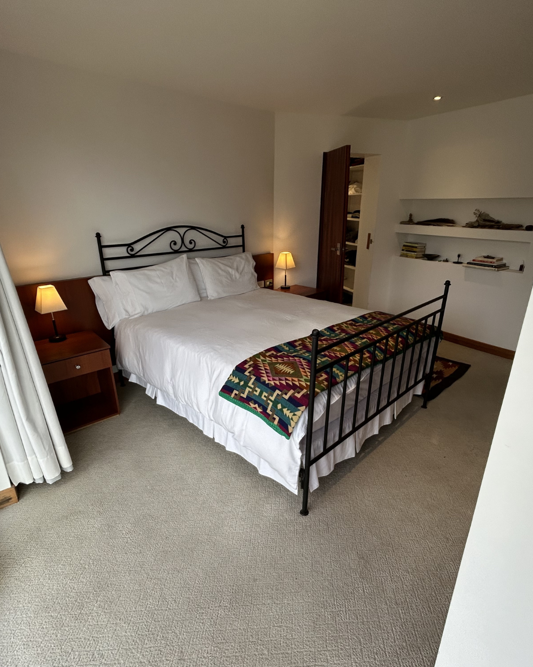 A neatly made bed with white linens and a colorful patterned blanket on top, flanked by two small nightstands with lamps, in a cozy, minimalist bedroom with built-in shelves and a partially open closet door.