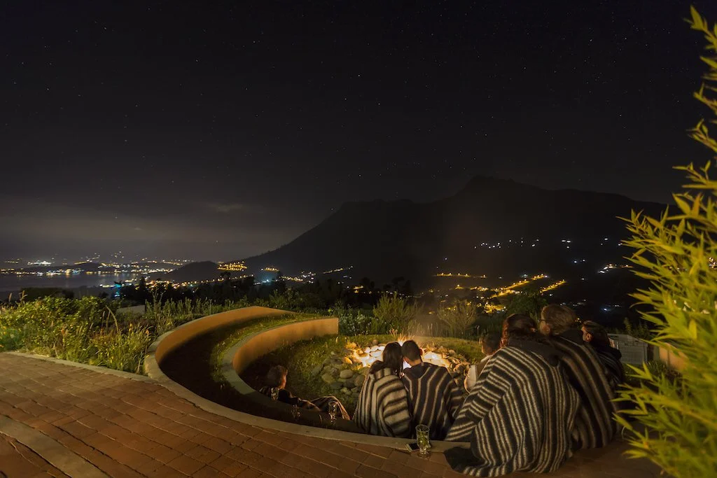 A group of people sitting around a fire pit at night, overlooking a city and mountain landscape with stars in the sky.