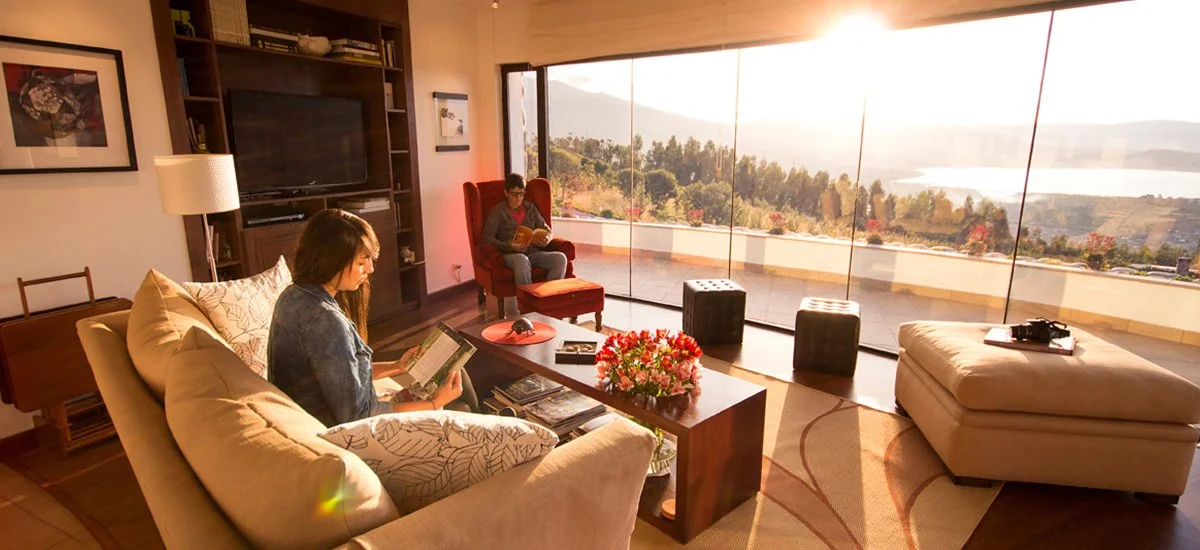 A living room with two people reading books, large glass sliding doors revealing a scenic outdoor view of trees and water, and modern furniture.
