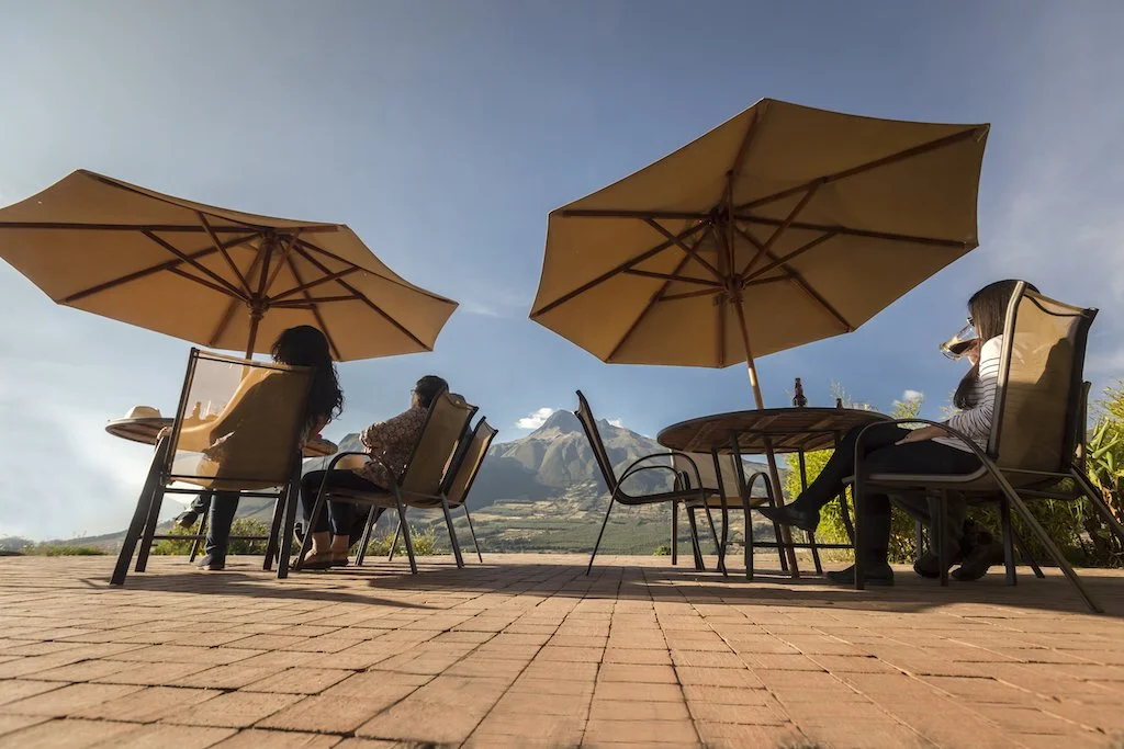 Three women sitting at outdoor tables with large umbrellas, enjoying a scenic view of mountains on a sunny day.
