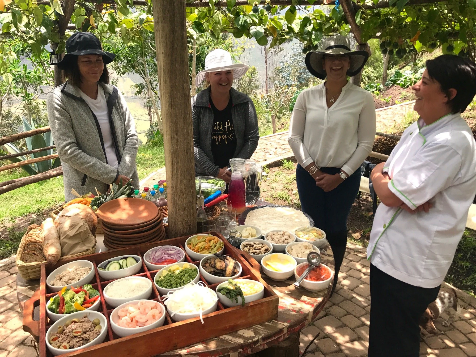 Women wearing sun hats and casual clothes gathered outdoors around a table filled with a variety of salads and food bowls, smiling and talking, with trees and greenery in the background.