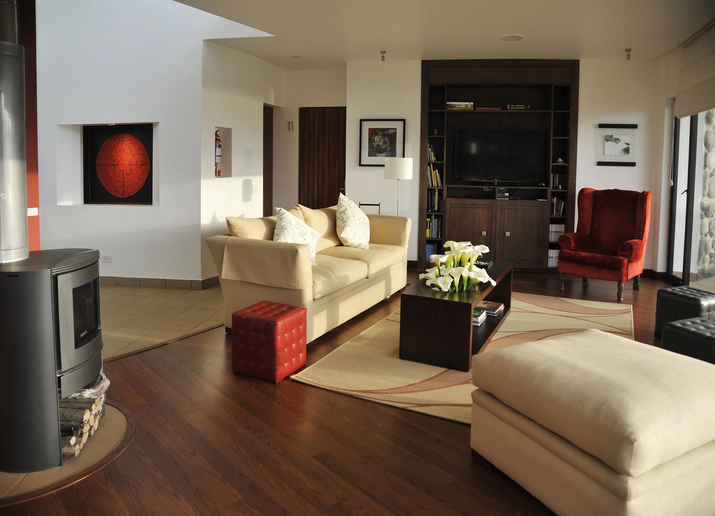 A living room with a white couch, red velvet armchair, dark wooden shelves with a television, decorated with pillows, a coffee table with a flower arrangement, and a wooden door in the background.