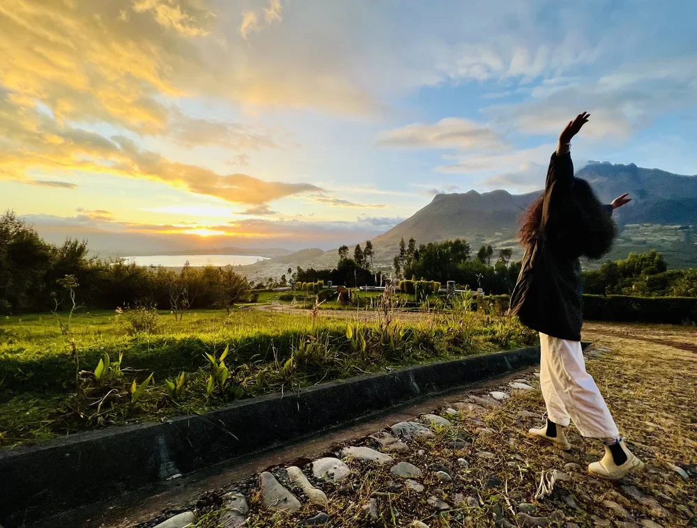 A woman with long hair standing on a trail with arms raised, overlooking a scenic landscape during sunset, with mountains, trees, and a body of water in the background.
