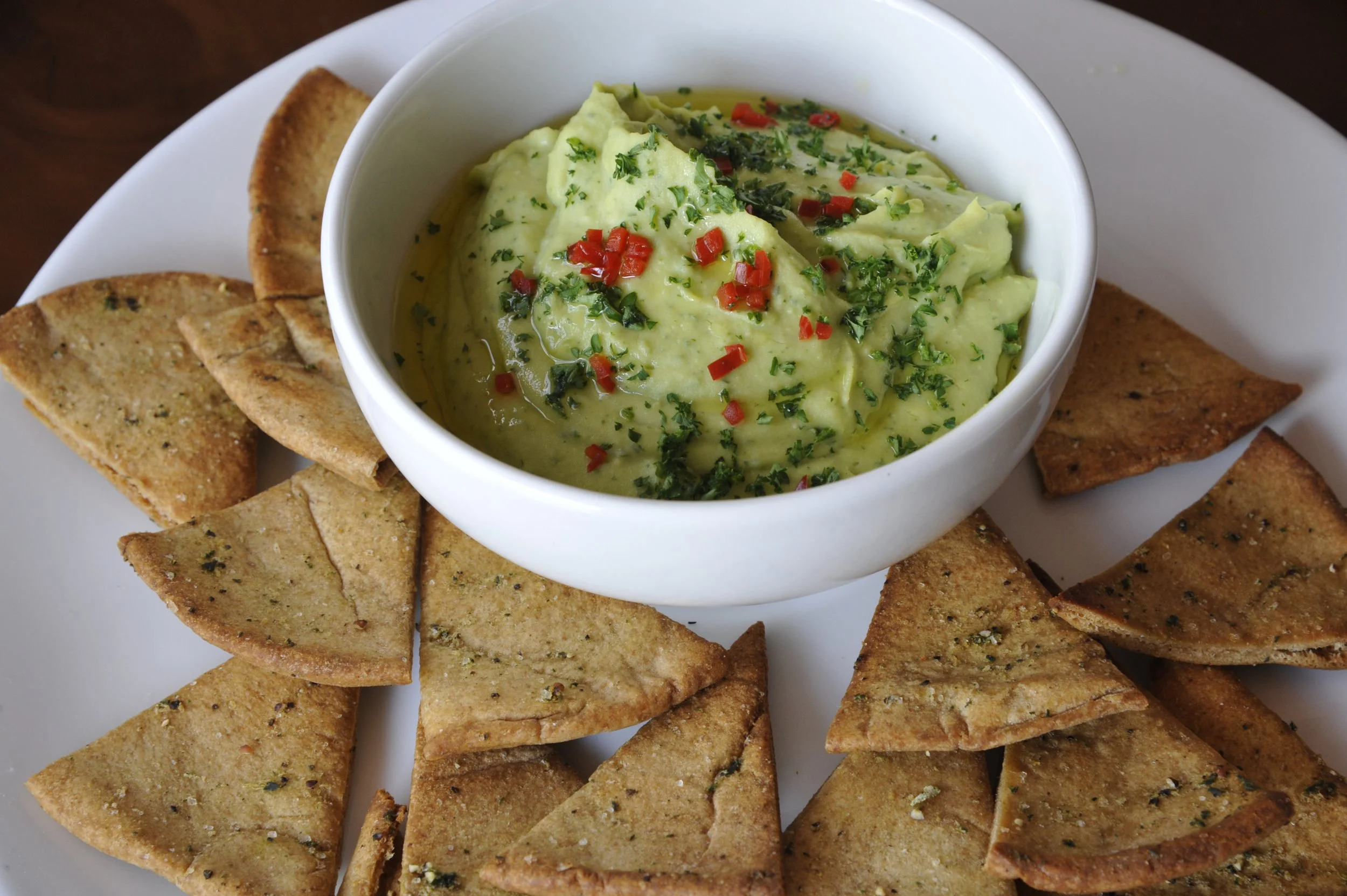 A white plate with triangles of garlic toast surrounding a bowl of guacamole topped with chopped red peppers and green herbs.