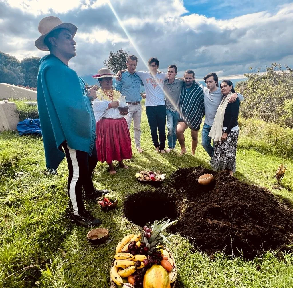 A group of people participating in a traditional outdoor ceremony, standing around a large hole in the ground with offerings including fruits, vegetables, and bowls placed nearby. Tall grass, trees, and a cloudy sky are in the background.