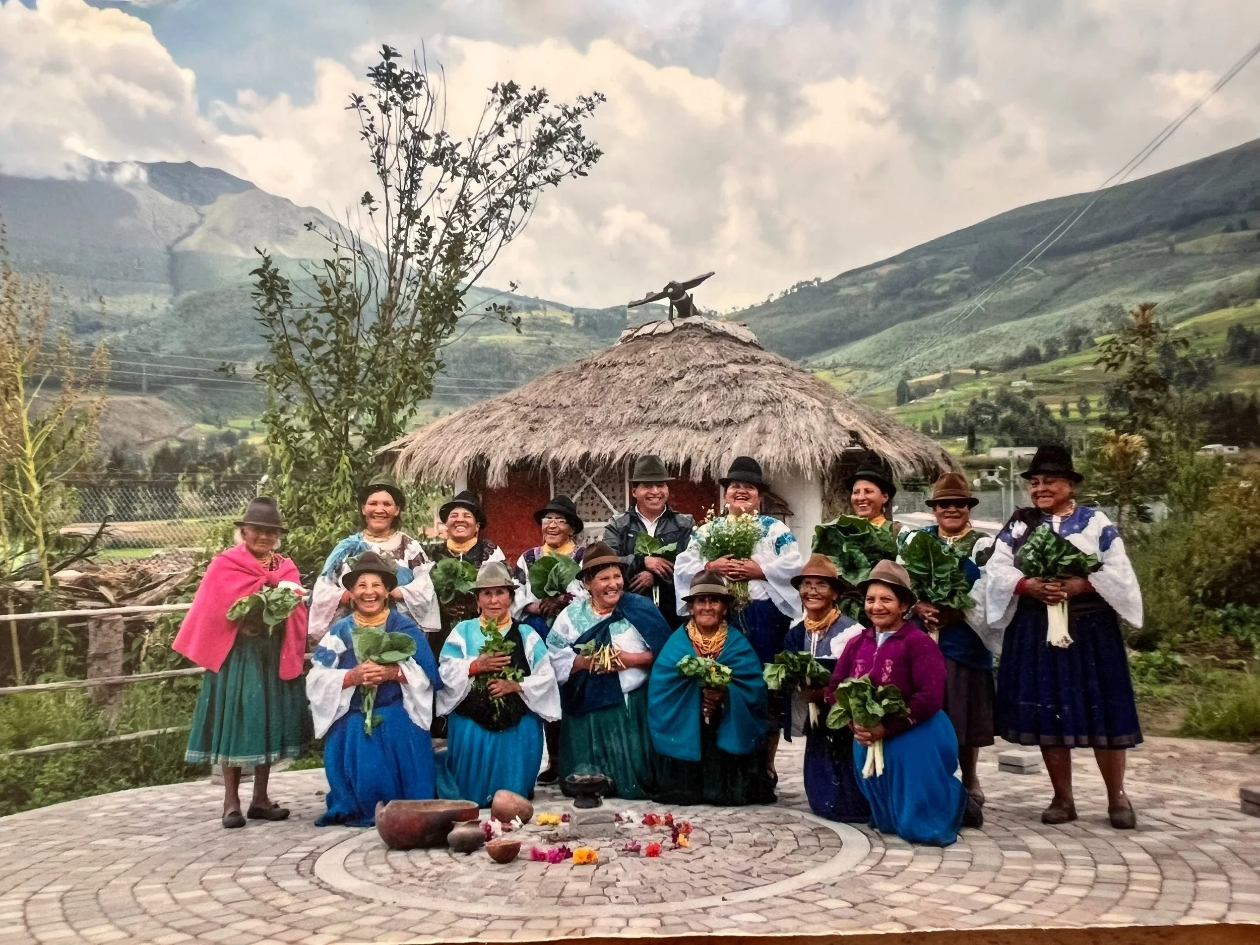 A group of people dressed in traditional clothing, some holding green plants, standing in front of a small round altar with flowers and offerings, outdoors in a mountainous area.