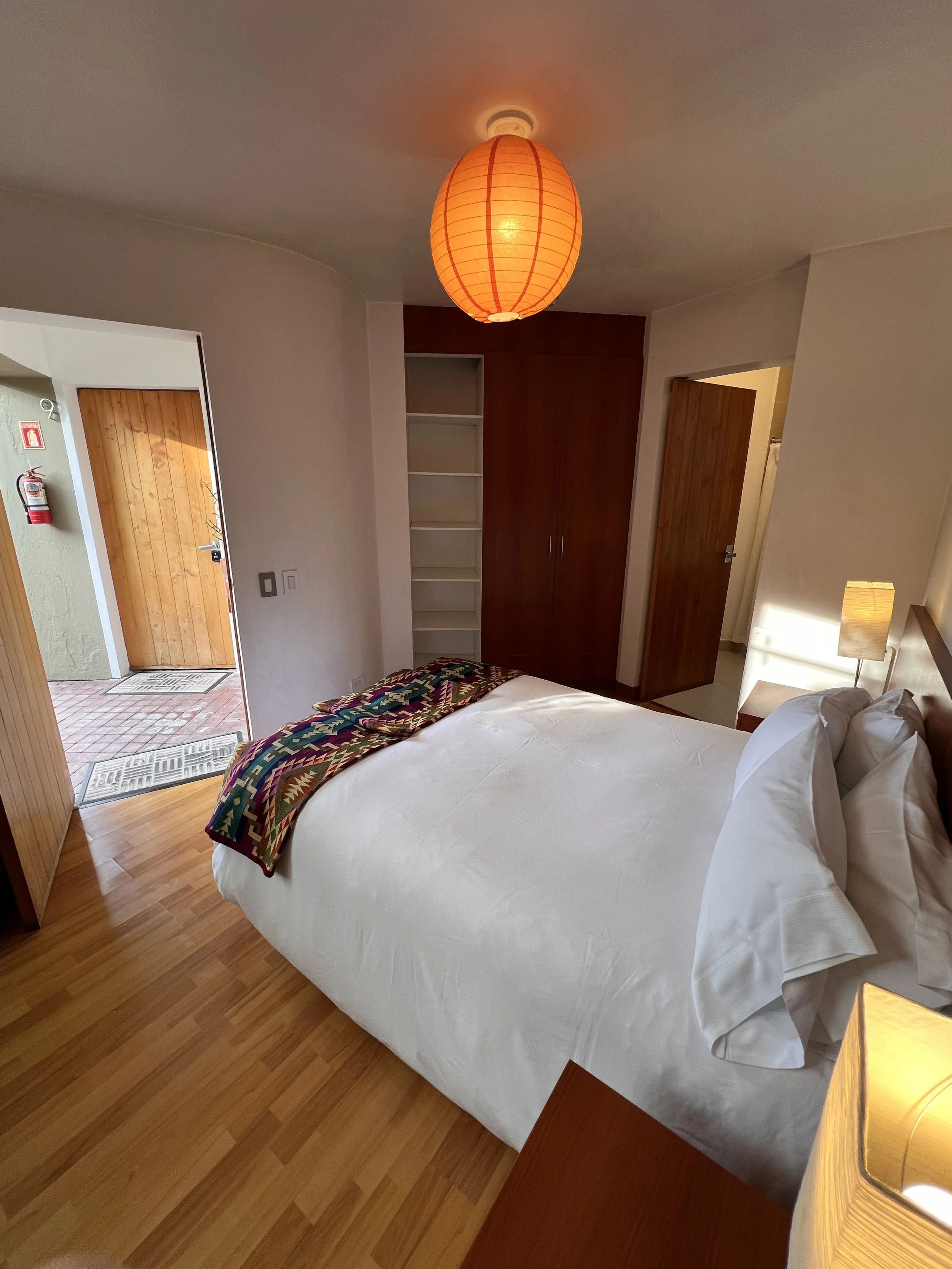 A hotel room with a bed, a lamp, a wooden wardrobe, and an entrance door visible, with a wooden floor and white walls.