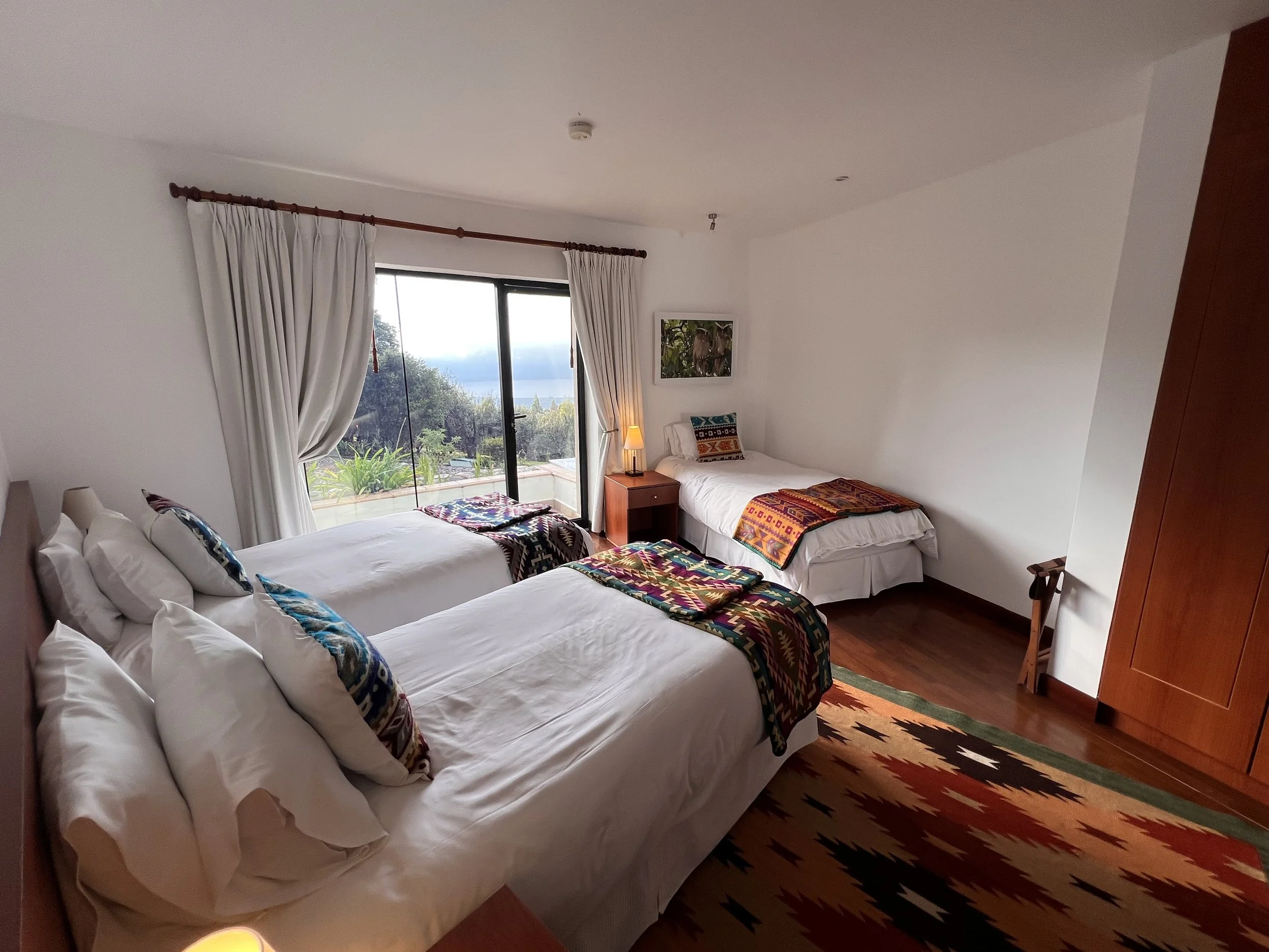 A bedroom with three beds, each covered with white sheets and colorful blankets, near a large window with white curtains, and a colorful rug on a wooden floor.