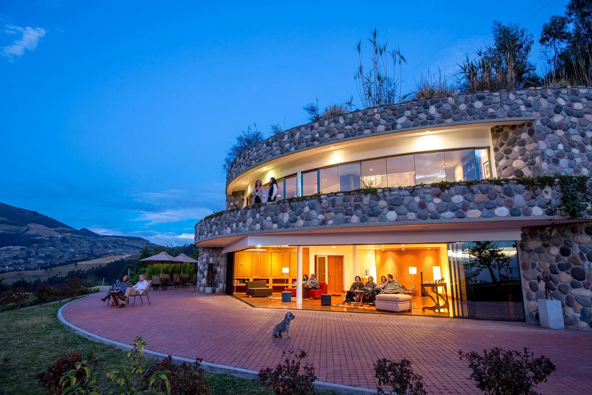 People relaxing and chatting outside a modern house with a stone facade during dusk, with a scenic mountain view in the background.