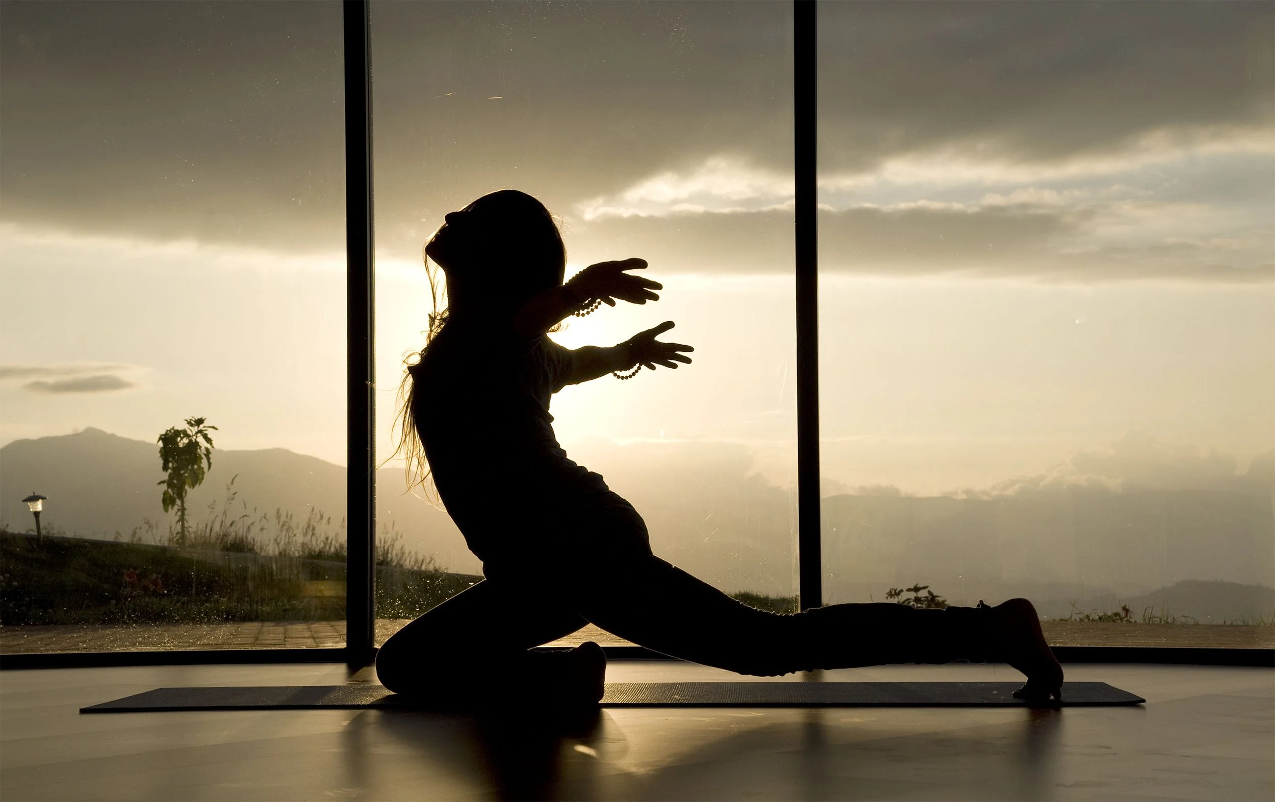 Silhouette of a woman practicing yoga indoors, sitting in a split pose, with large windows showing an outdoor landscape and cloudy sky at sunset.